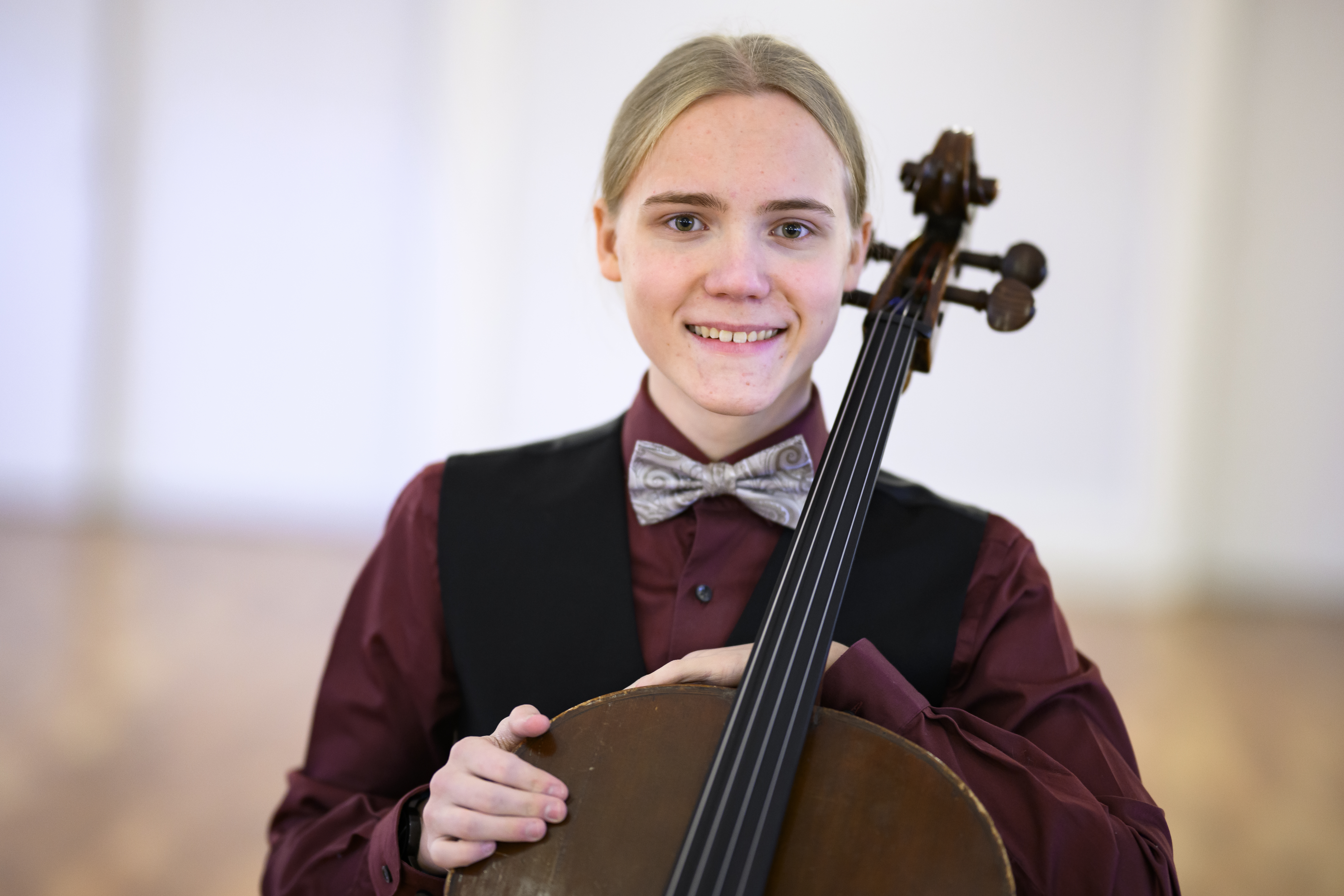 En ung kille i fluga och väst sitter med en cello lutad mot sig. Han ler stort.