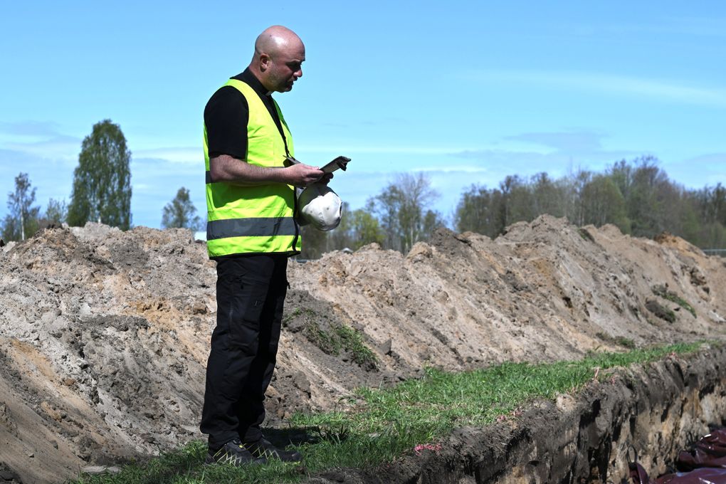 En man i gul varselväst och svar prästskjorta står med keps i handen och sänkt huvud. han står på en gräsplätt bland jordhögar och gropar på en tillfällig begravningsplats.