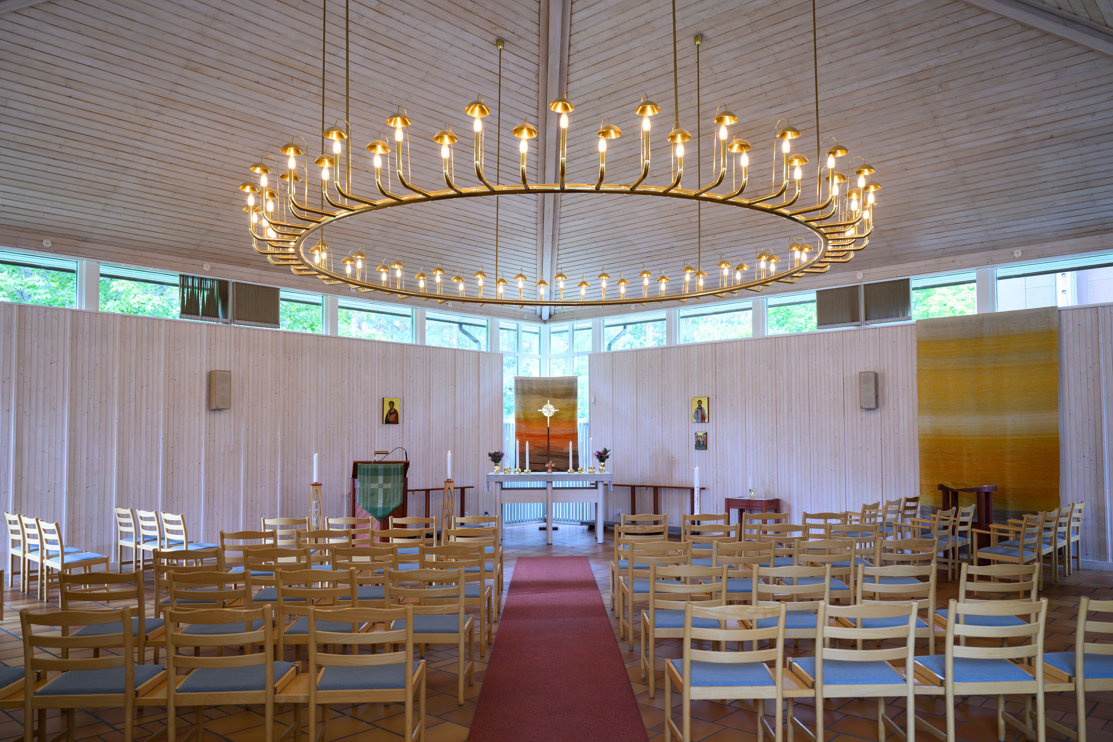 Interiör från S:t Lars kyrka. Ljusa väggar och tak, med smala ljusinsläpp högt upp. Möblerat med lösa stolar på var sida mittgången. I taket en stor rund ljuskrona.
