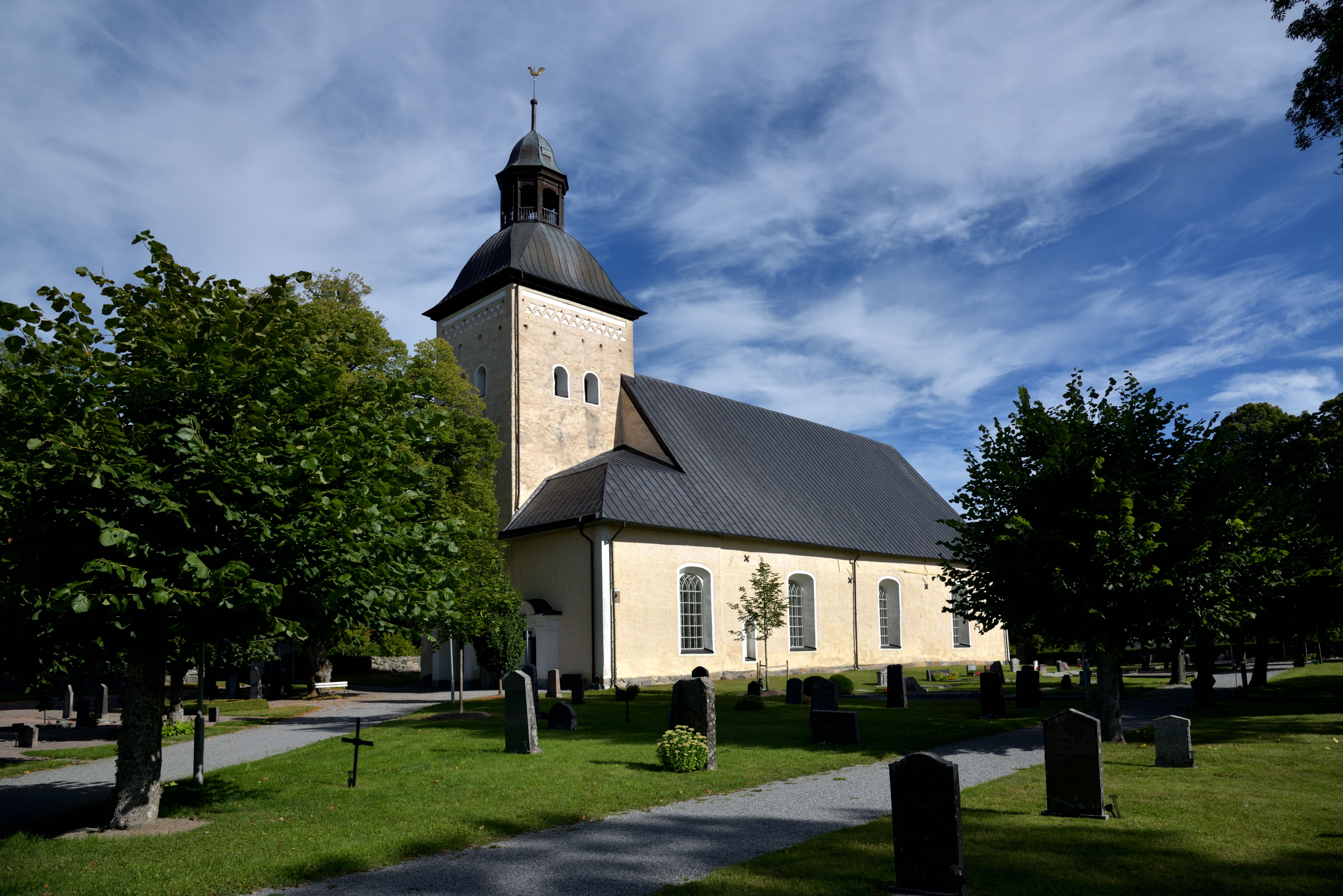 En gulaktig kyrka med ett torn. Gröna lövträd står på kyrkogården runt kyrkan. Himlen är blå.