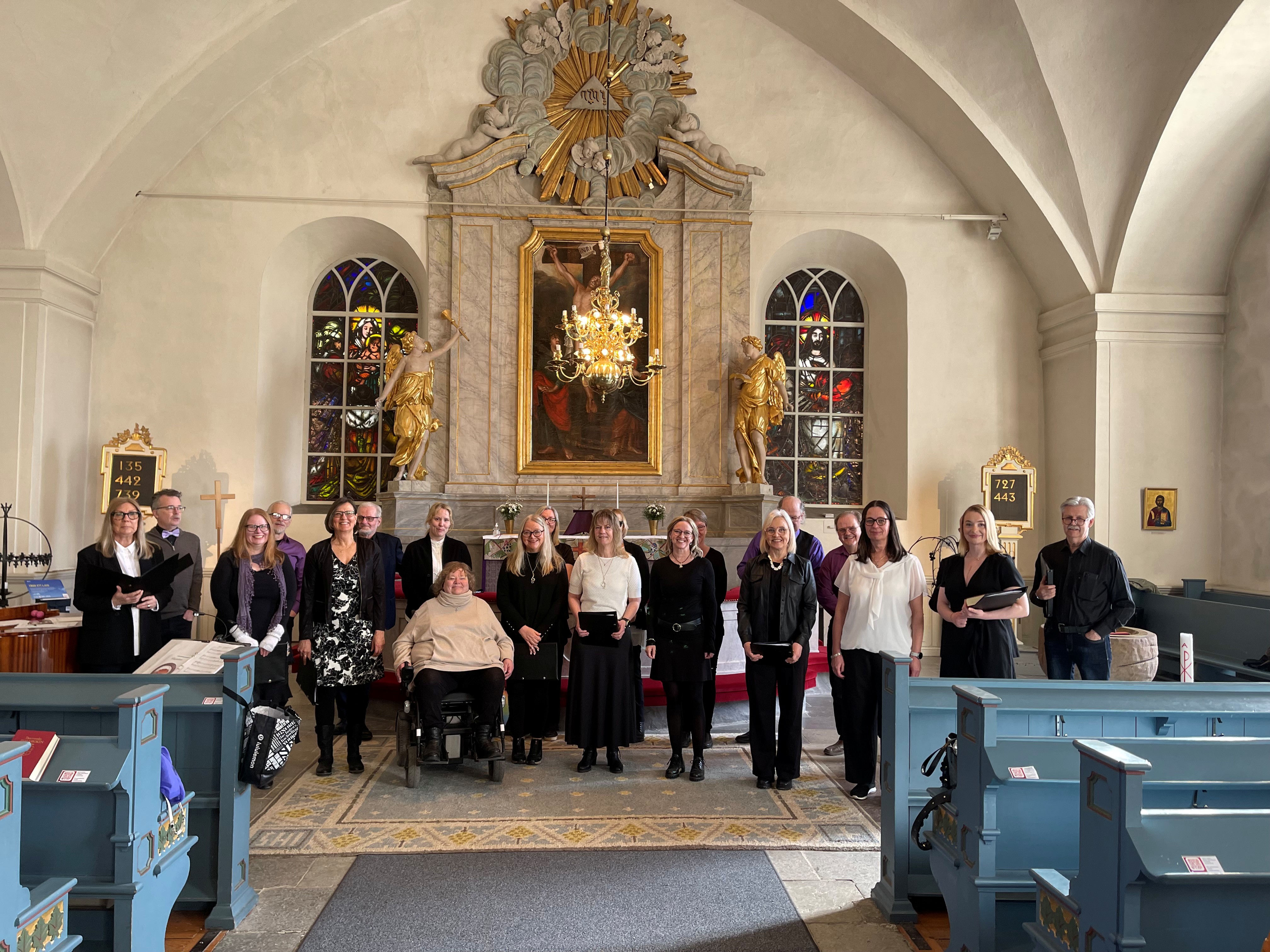 En kör med människor i blandade åldrar, olika kön och funktion står uppställda i koret i en kyrka.
