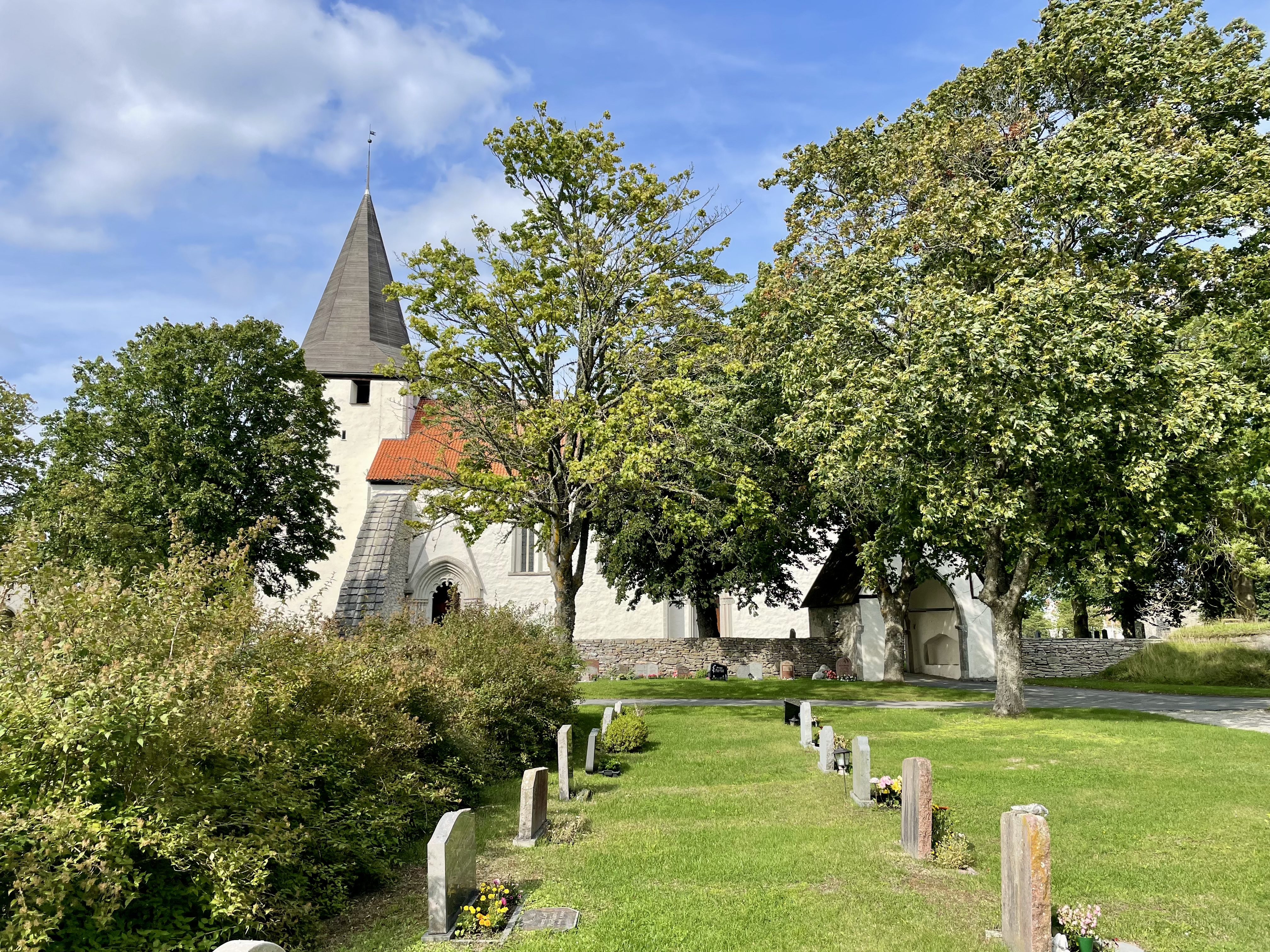 Bunge kyrka från söder, man ser gravstenar och lummiga träd och gräsmatta i förgrunden