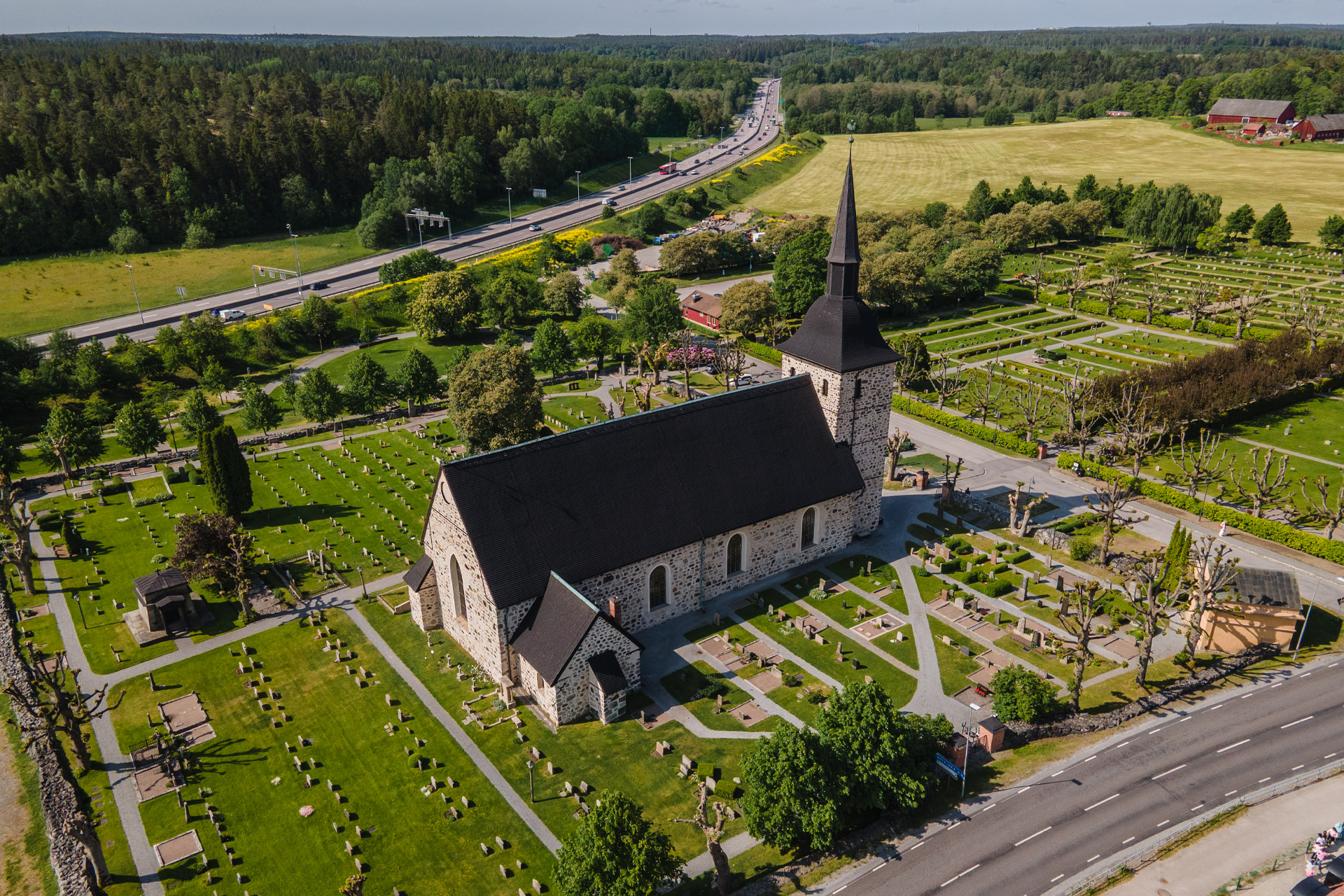 Botkyrka kyrka fotograferad från ovan. Runt kyrkan syns kyrkogården, grönskande träd och gräs och en motorväg.
