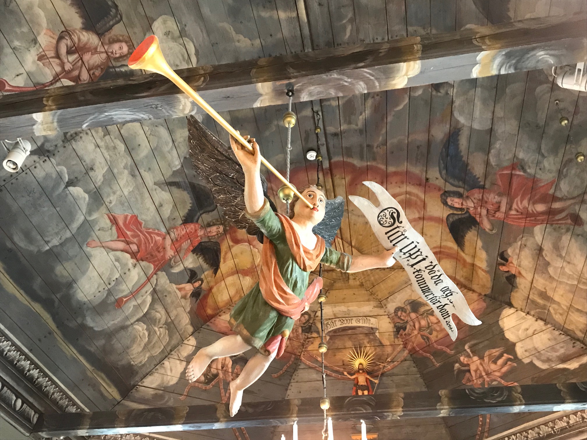 En träängel hänger i ett målat kyrktak. Ängeln blåser i ett stort horn och har ett banner med text i handen. 