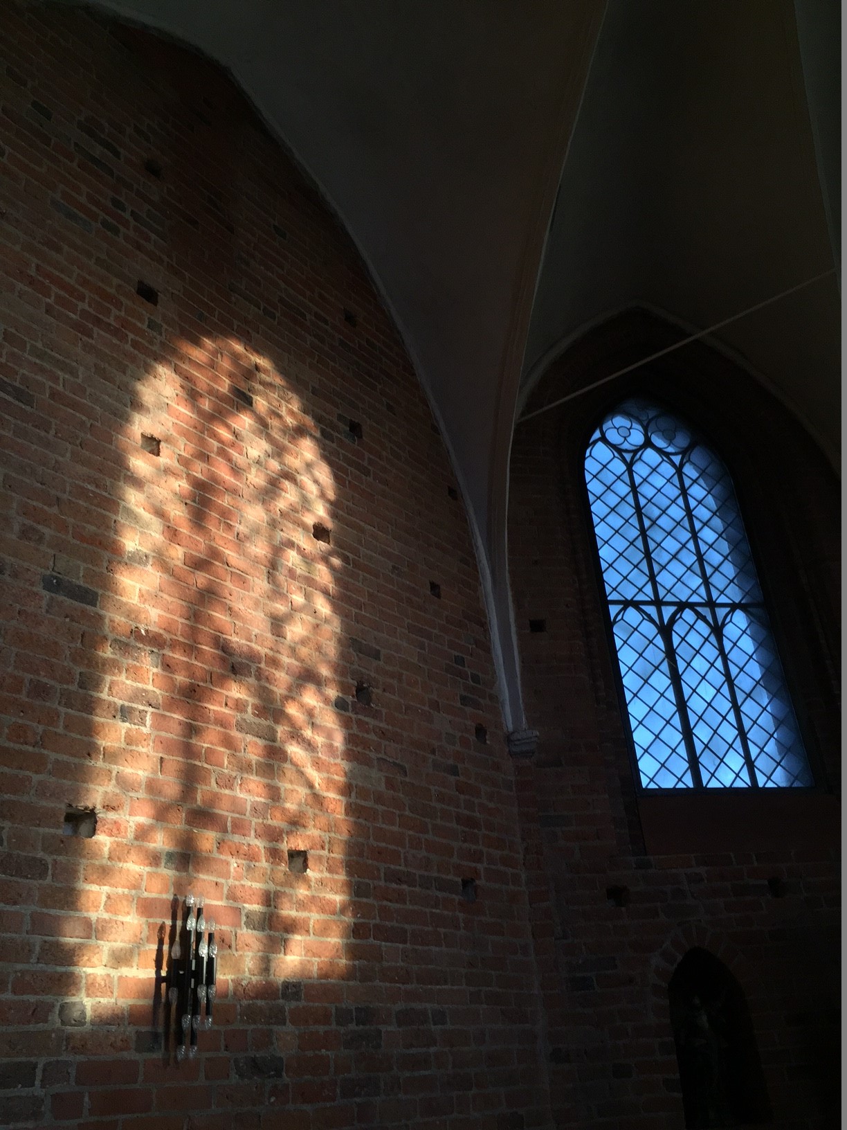 Ett av Klosterkyrkans korfönster med spröjs syns till höger i bilden. Till vänster syns en spegling på tegelväggen av ett av de andra fönstren.  