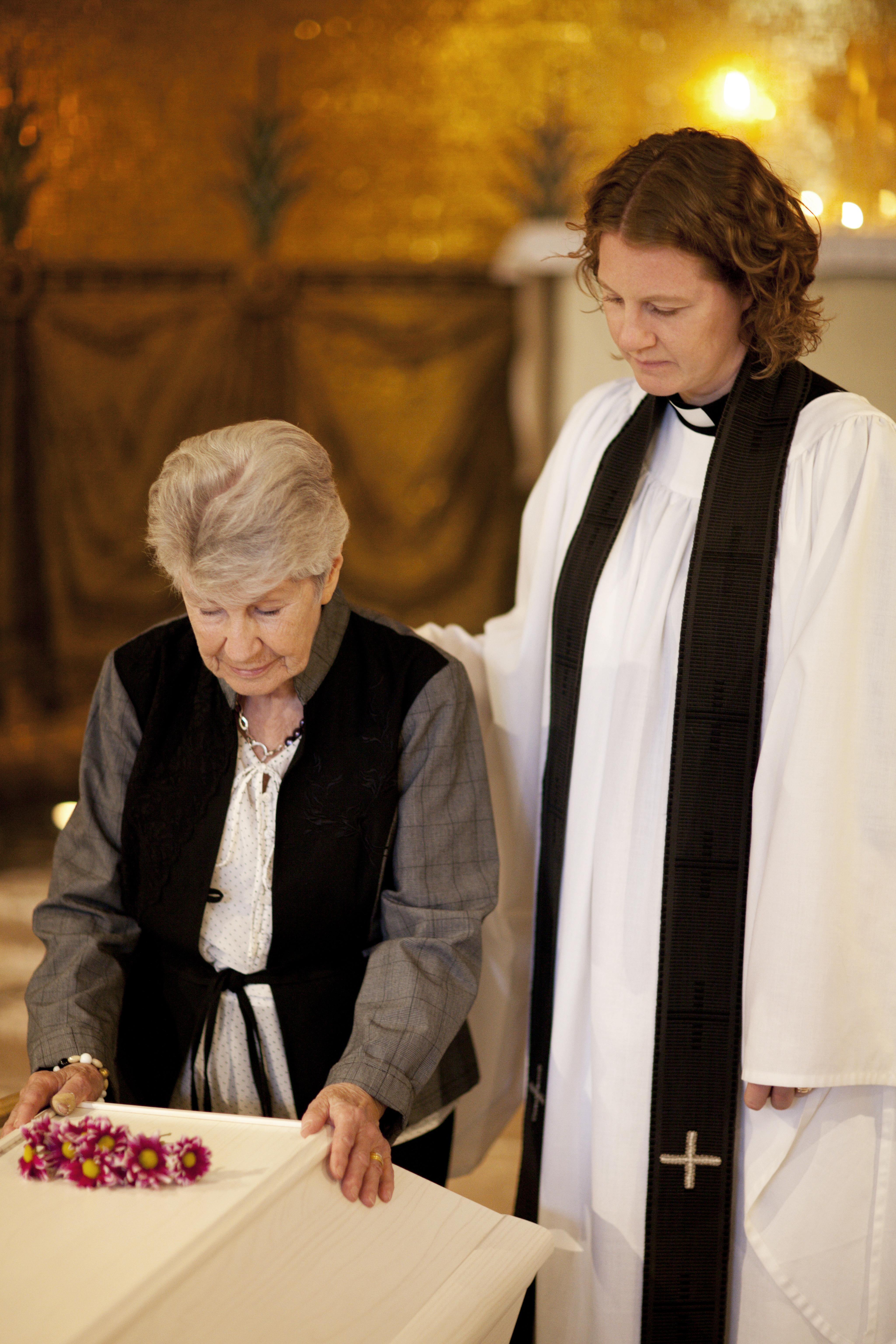 En äldre kvinna har lagt sina händer på en kista. En kvinnlig präst håller handen på hennes rygg.