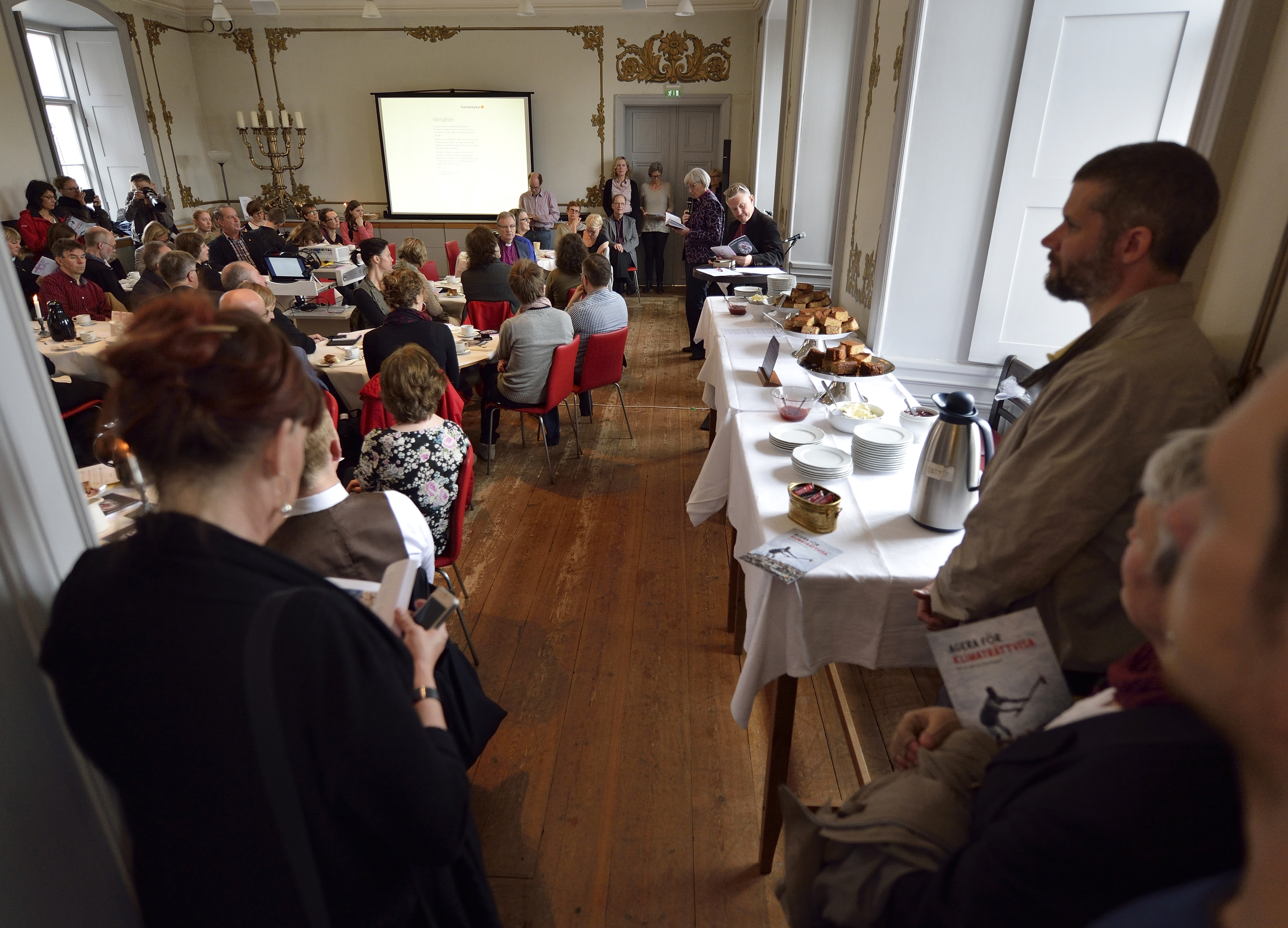 En grupp människor sitter i ett rum och lyssnar på en föreläsning och fikar.