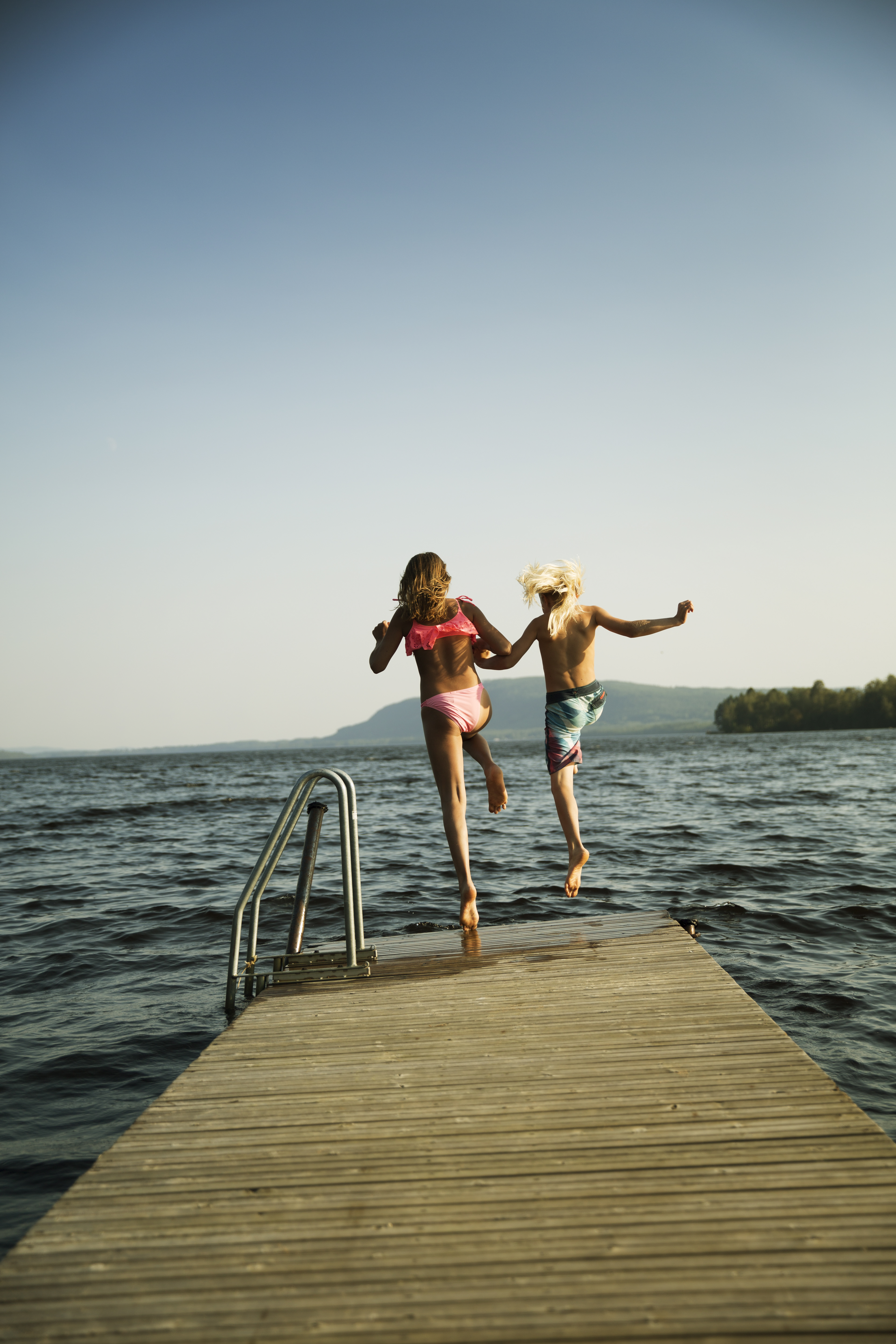 Två barn håller hand och hoppar från bryggan i vattnet.