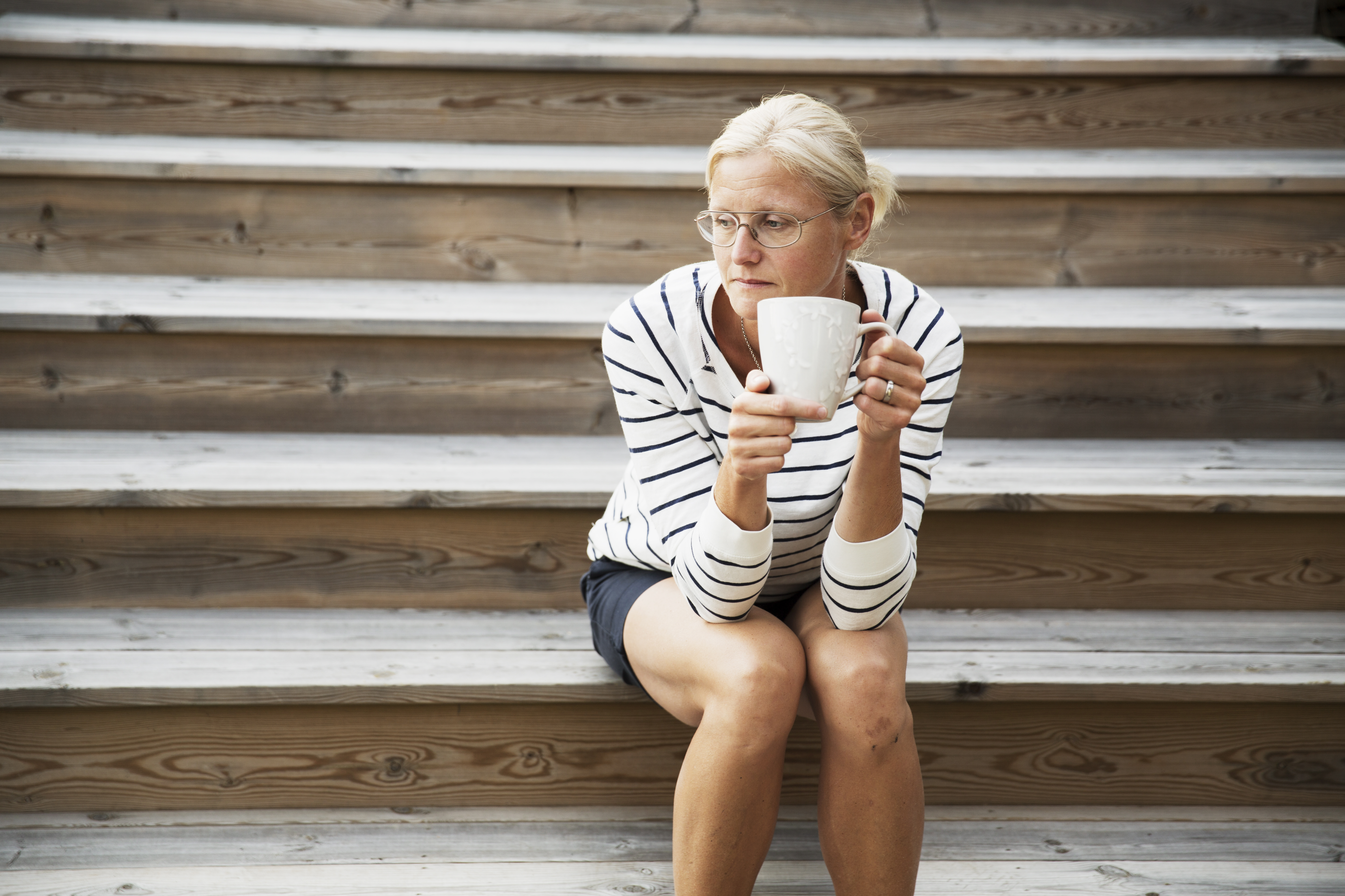 En kvinna sitter på trätrappa med en kaffekopp i handen.