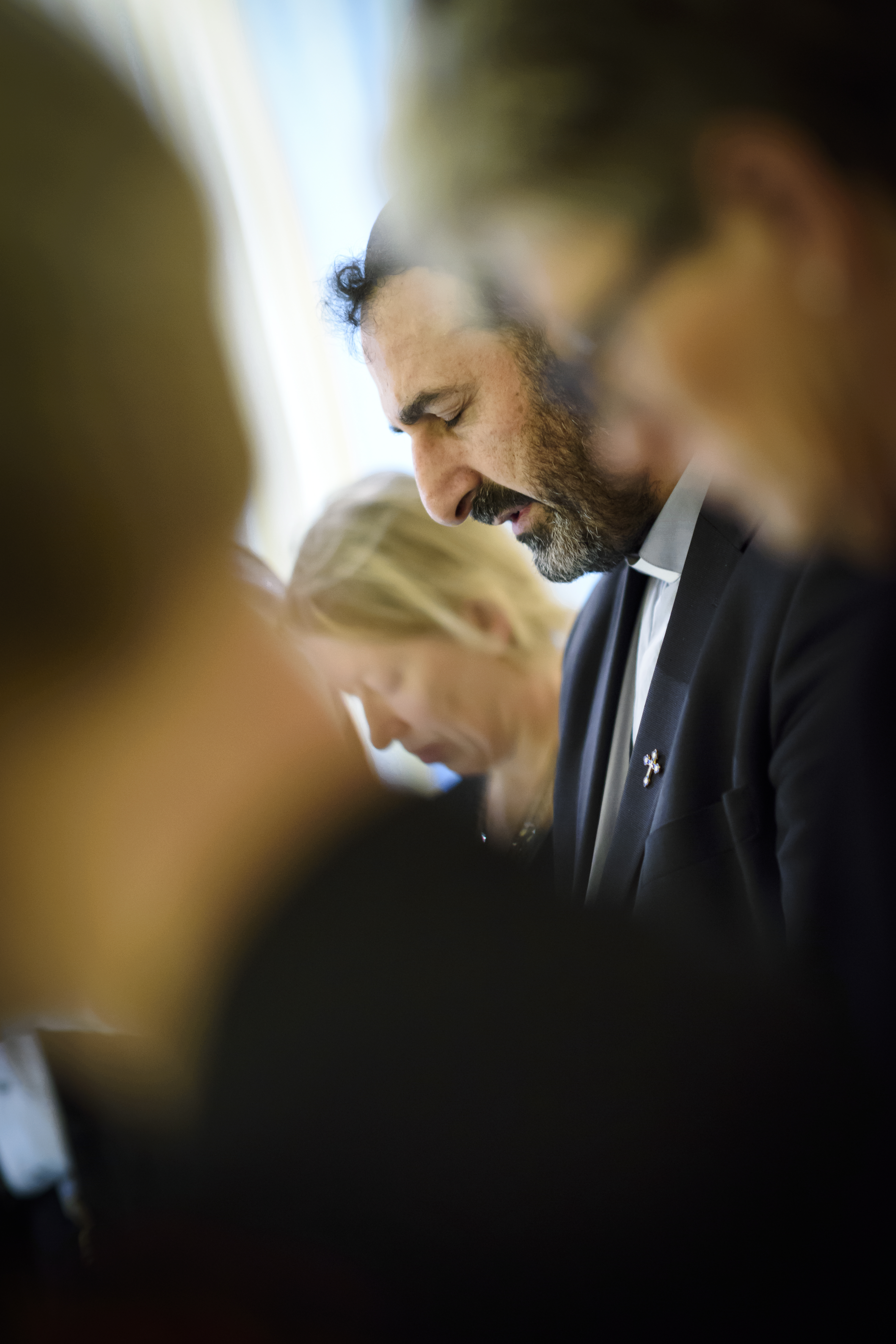 En man i kostym står och blundar med huvudet framåtlutat. Suddiga personer i förgrunden.