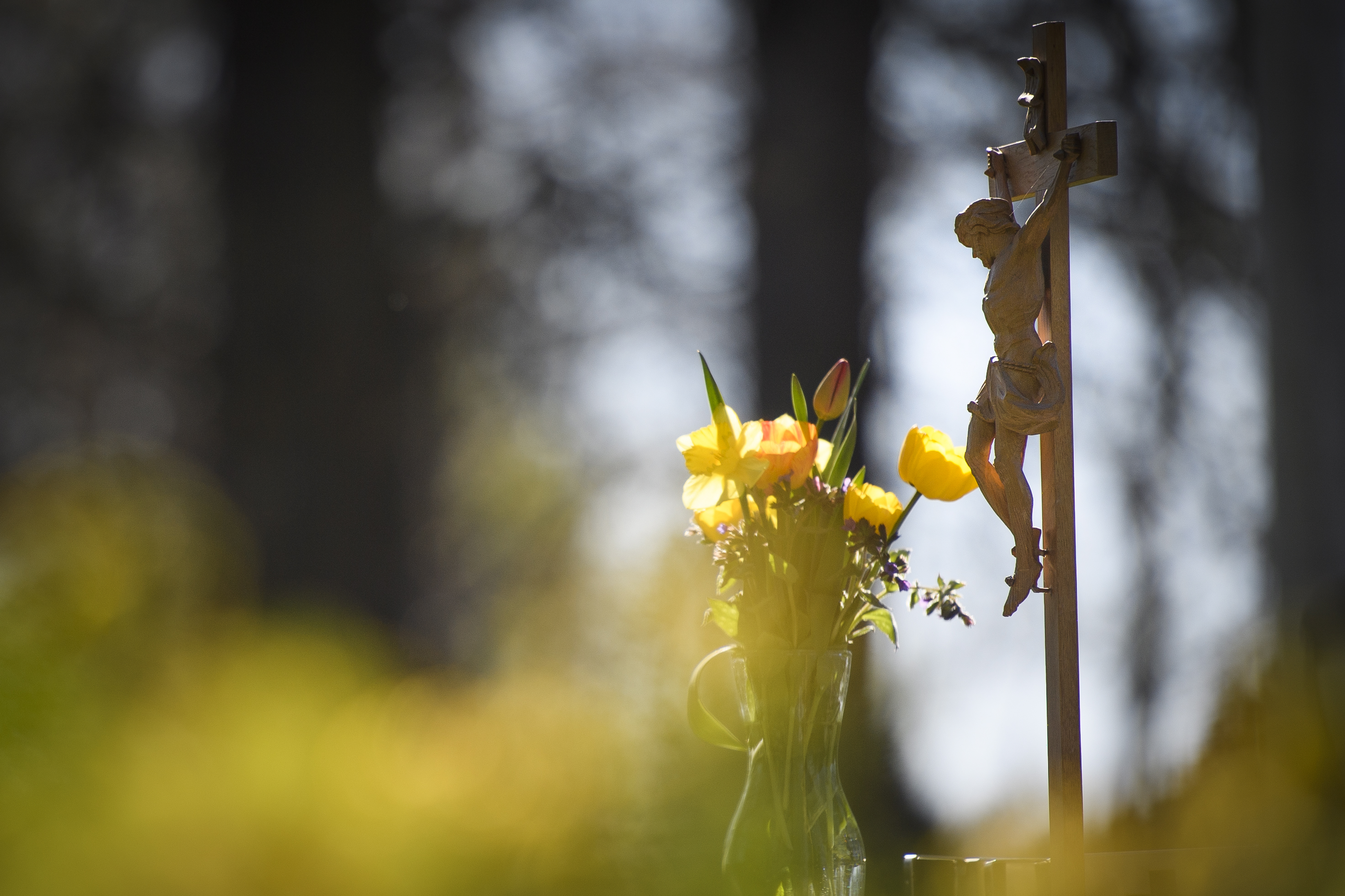 Ett krucifix i trä och en vas med gula blommor står utomhus.