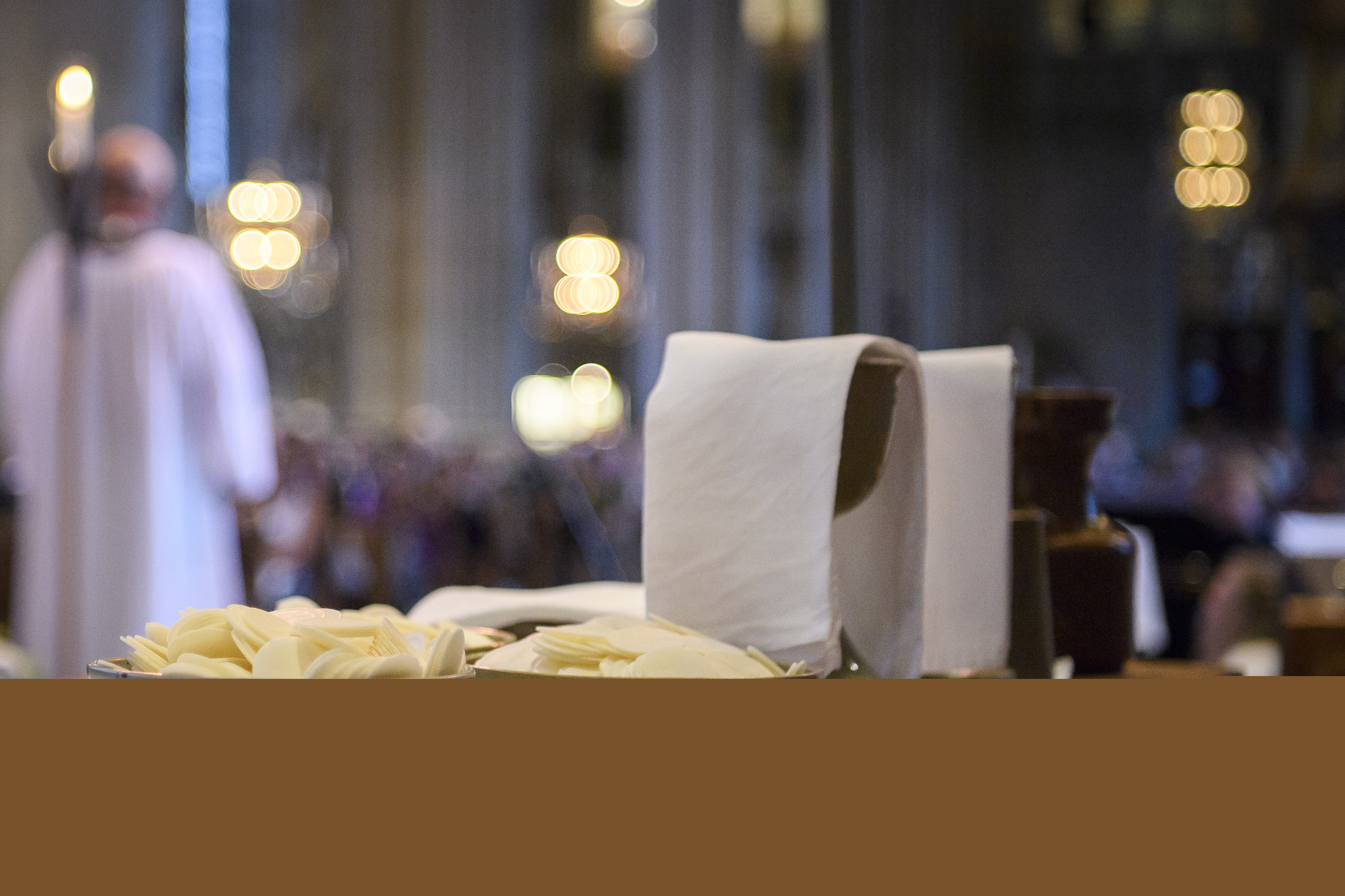 Nattvardsvin och oblat står uppdukat på ett bord. En präst och en många kyrkobesökare skymtas i bakgrunden.