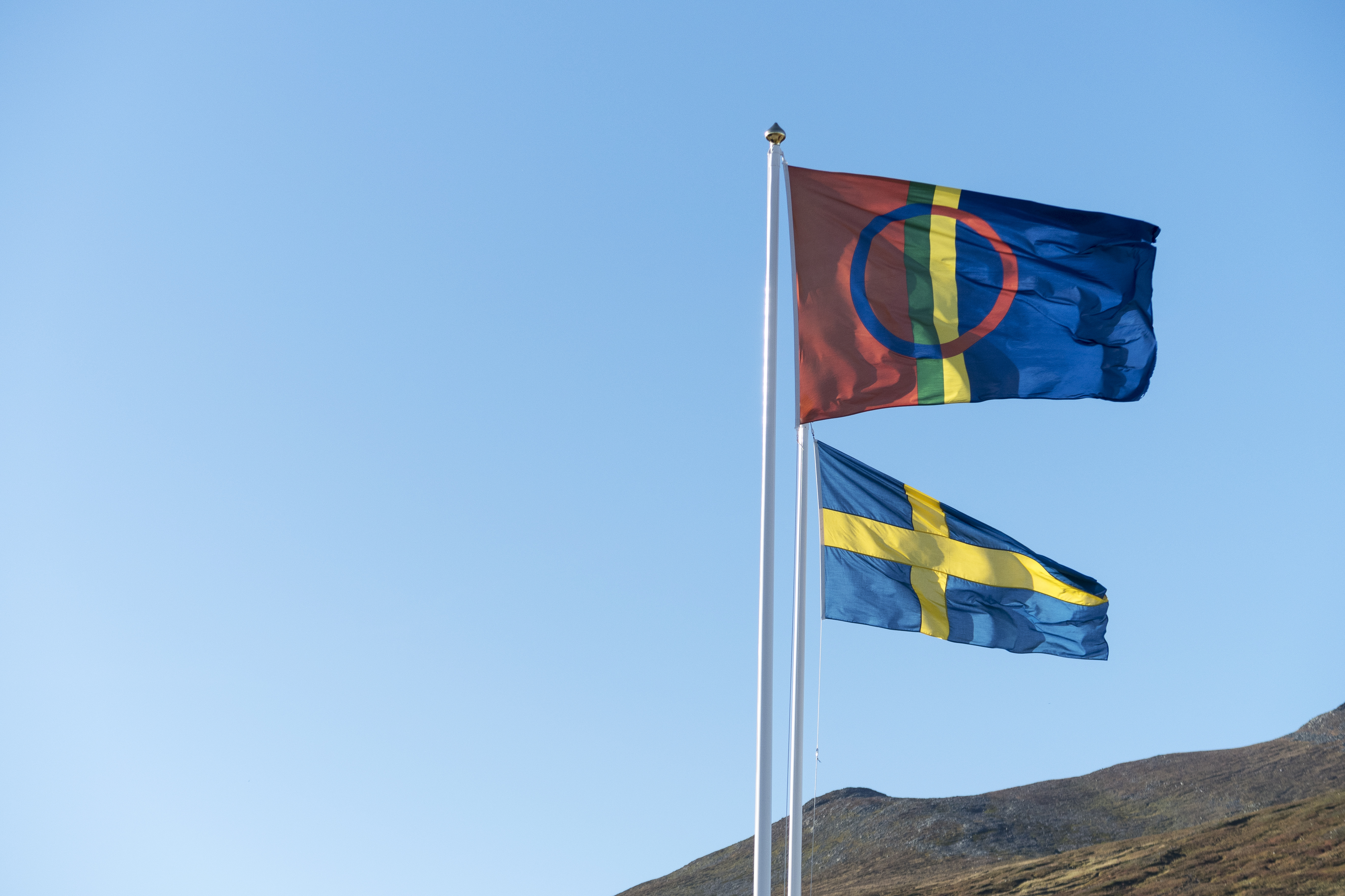 Två flaggstänger med samiska och svenska flaggan med blå himmel och berg i bakgrunden.