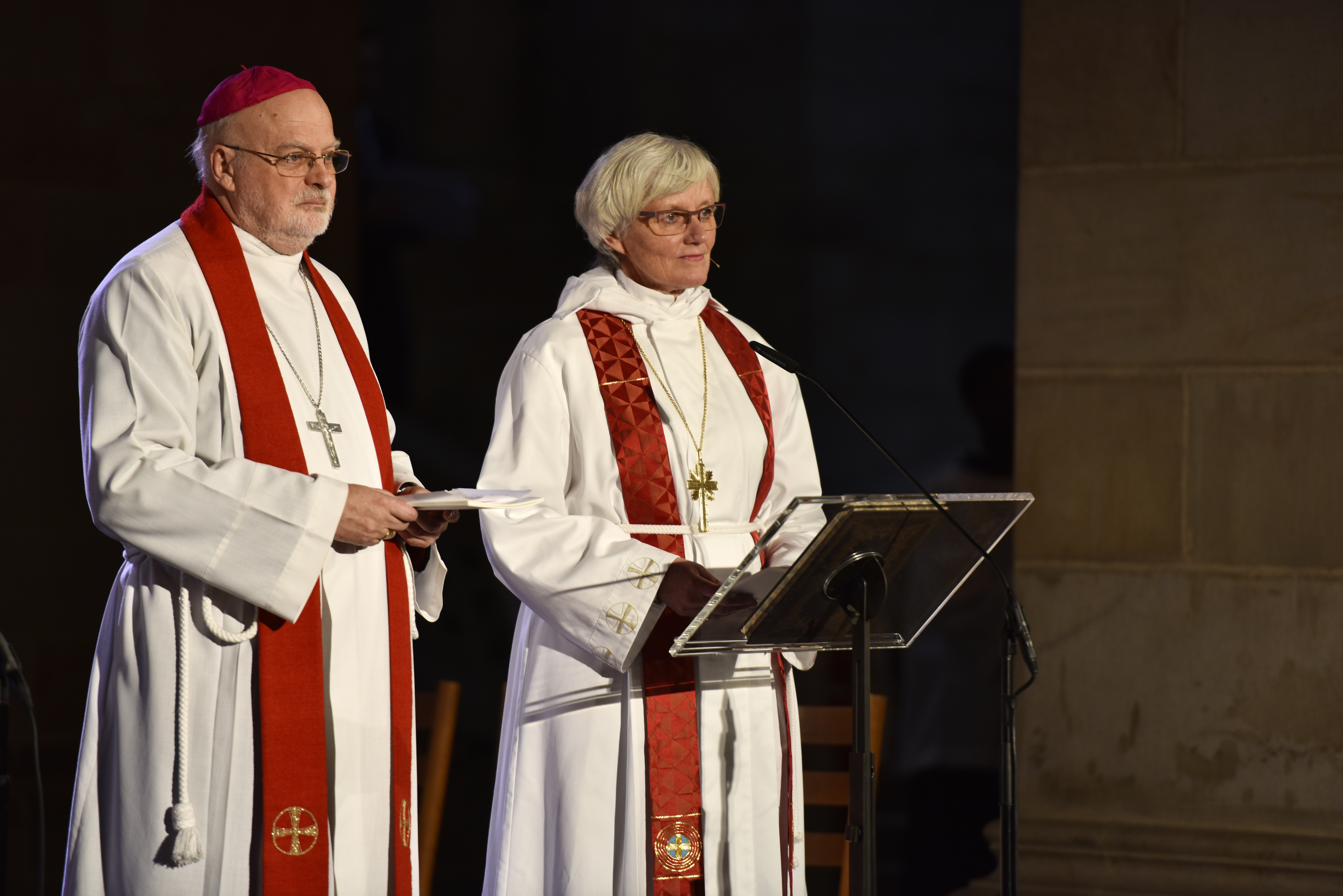 Biskoparna Anders Arborelius och Antje Jackelén står i strålkastarljus i en kyrka och talar i en mikrofon.