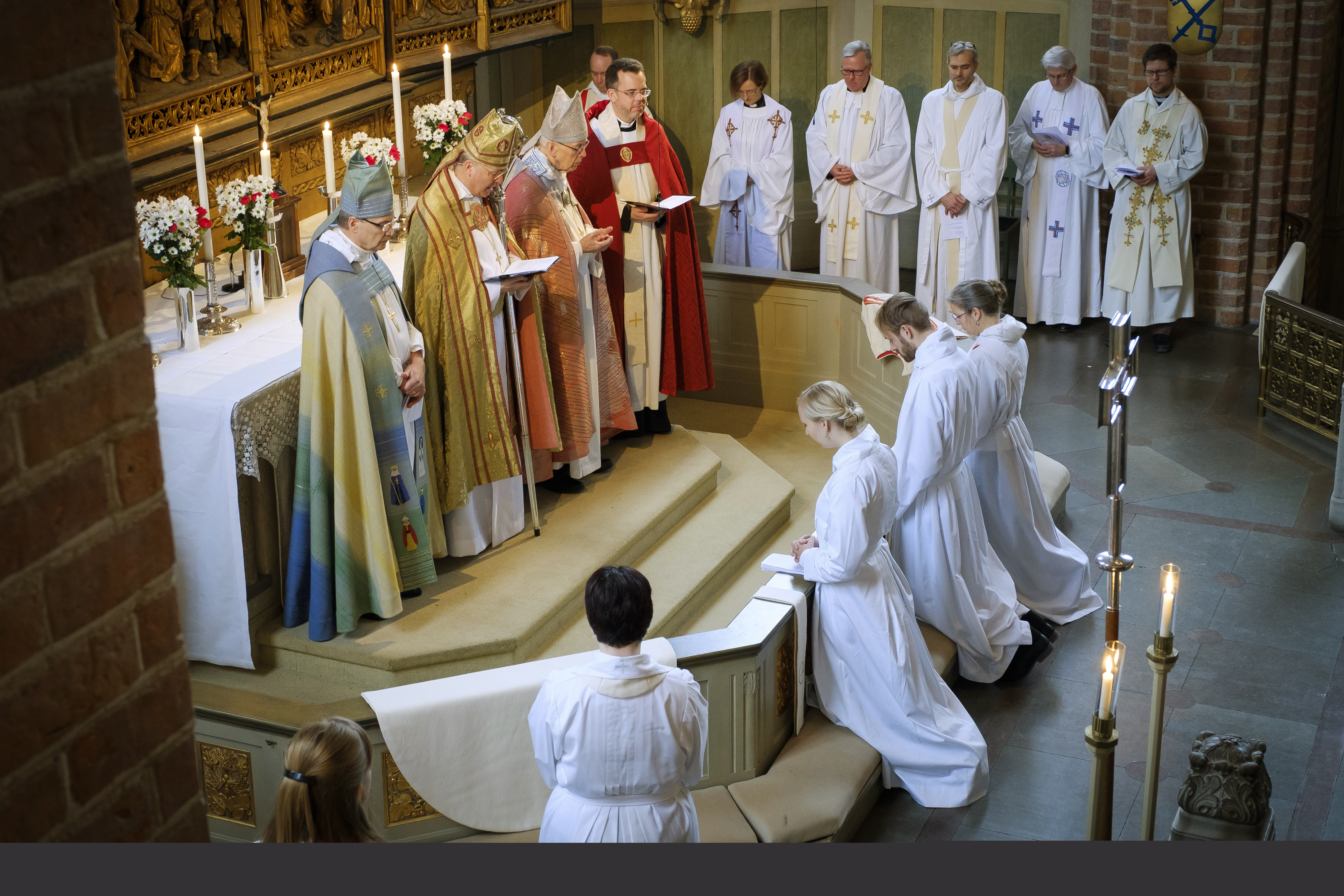 Fyra biskopar står vid ett altare, vända mot tre vitskrudade personer som knäböjer.