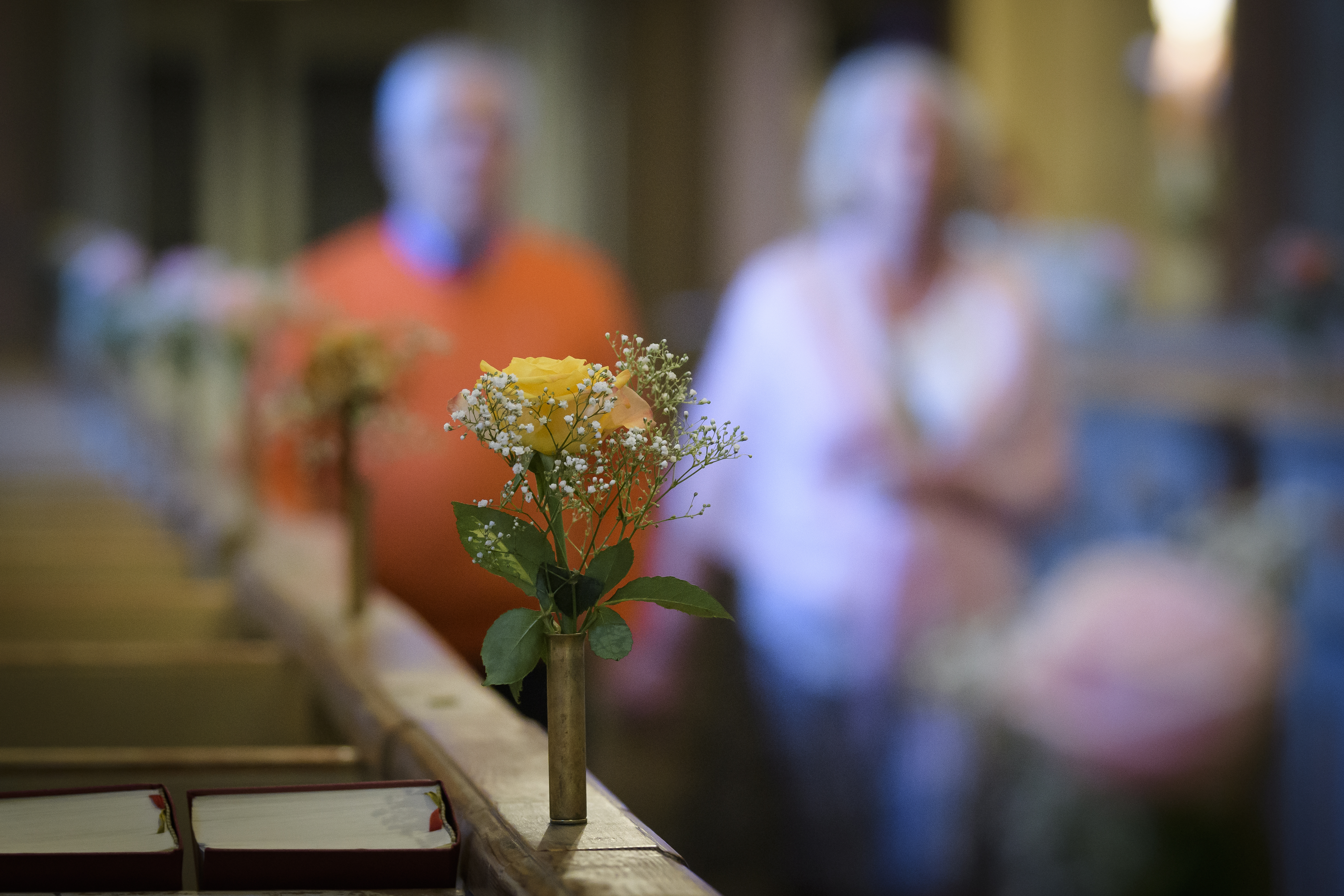 Små vaser med rosor står på bänkraderna i en kyrka. En man och en kvinna syns suddigt gå hand i hand i mittgången.