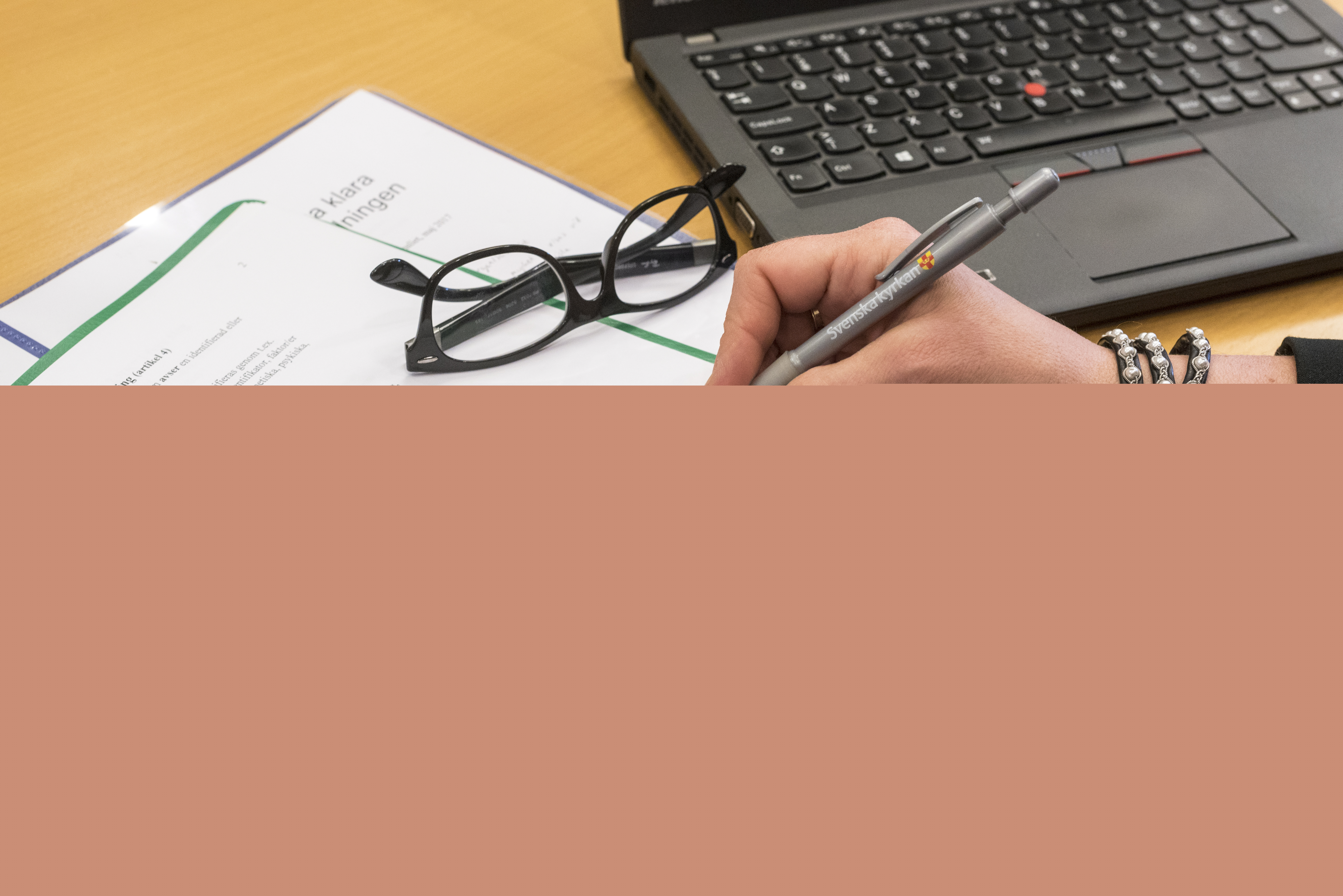En person med en penna i handen sitter med ett texthäfte och en dator framför sig.