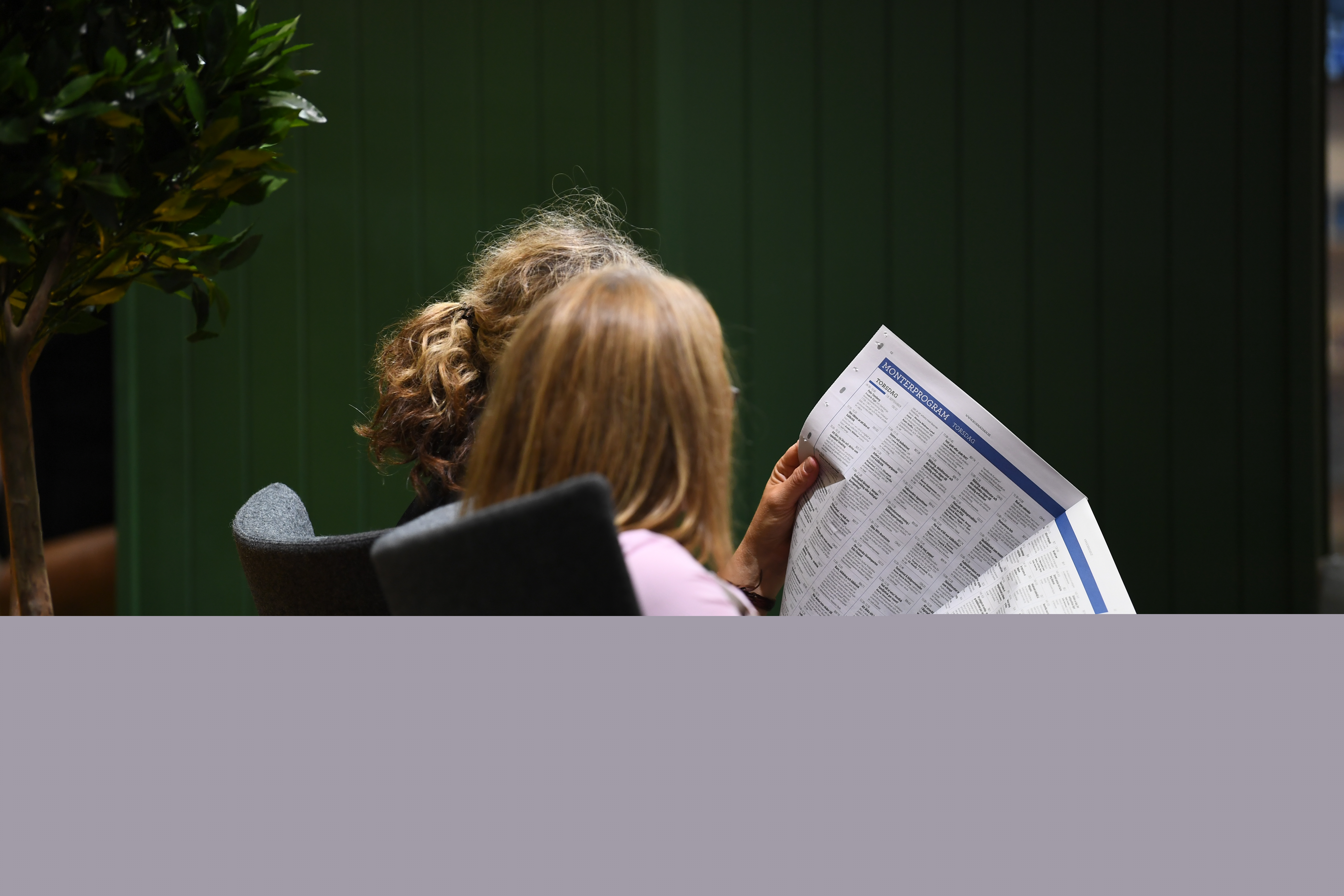 Två kvinnor sitter bredvid varandra inomhus och läser en tidning.