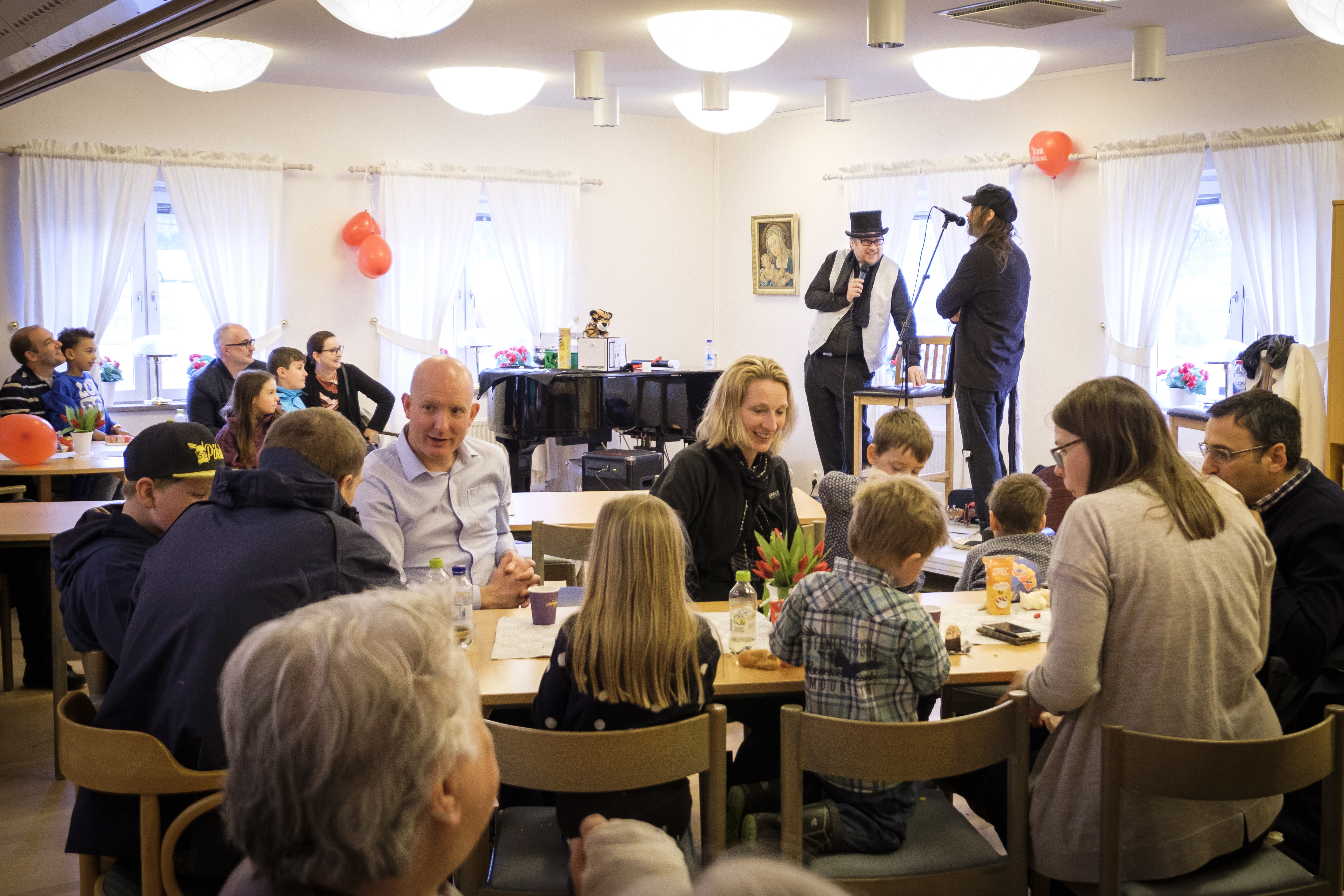 En grupp vuxna och barn sitter i en sal och fikar. I bakgrunden står två musiker på en liten scen.