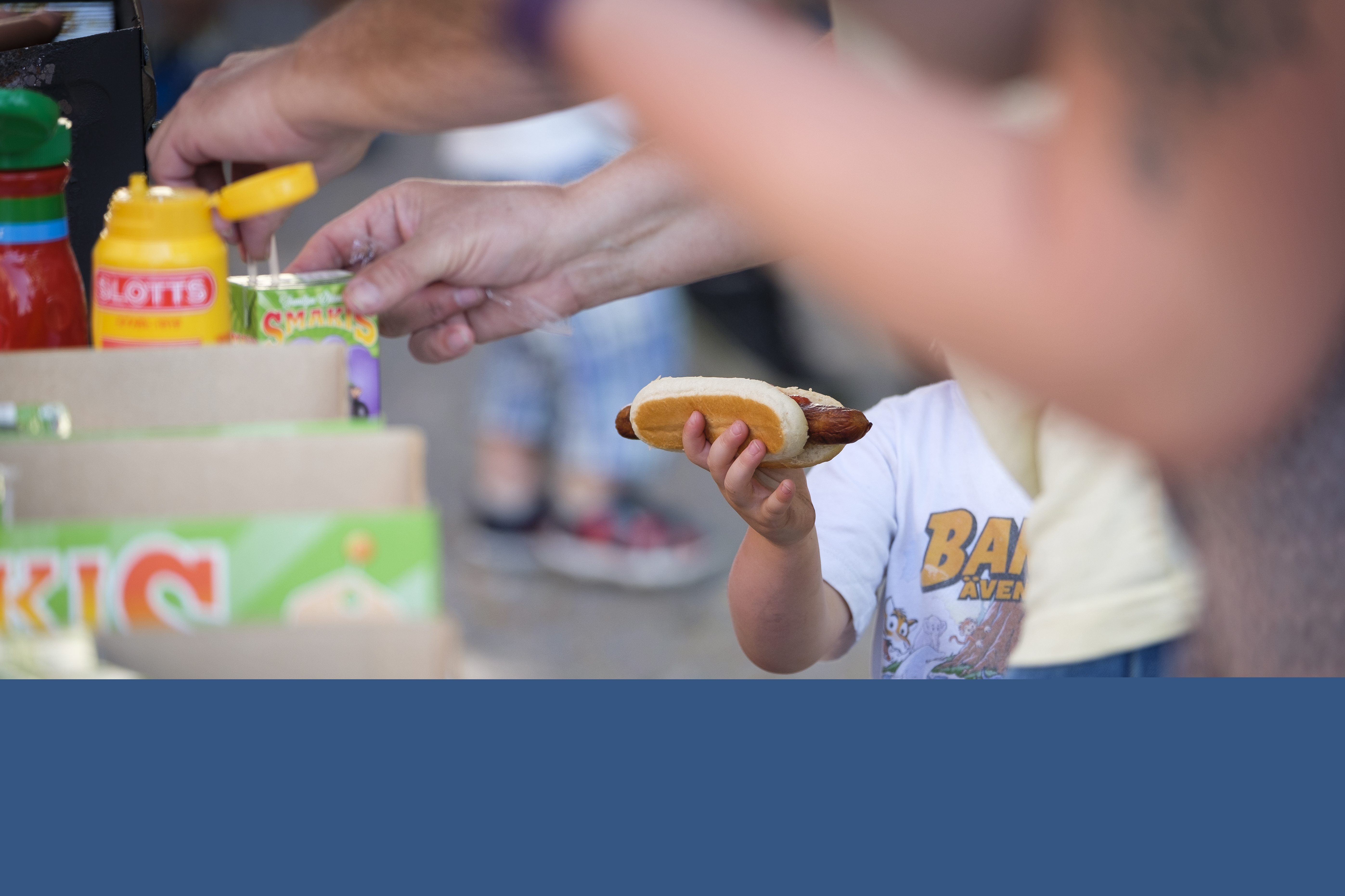 Ett barn står vid ett korvstånd med en varmkorv i handen. Suddiga personer i förgrunden.