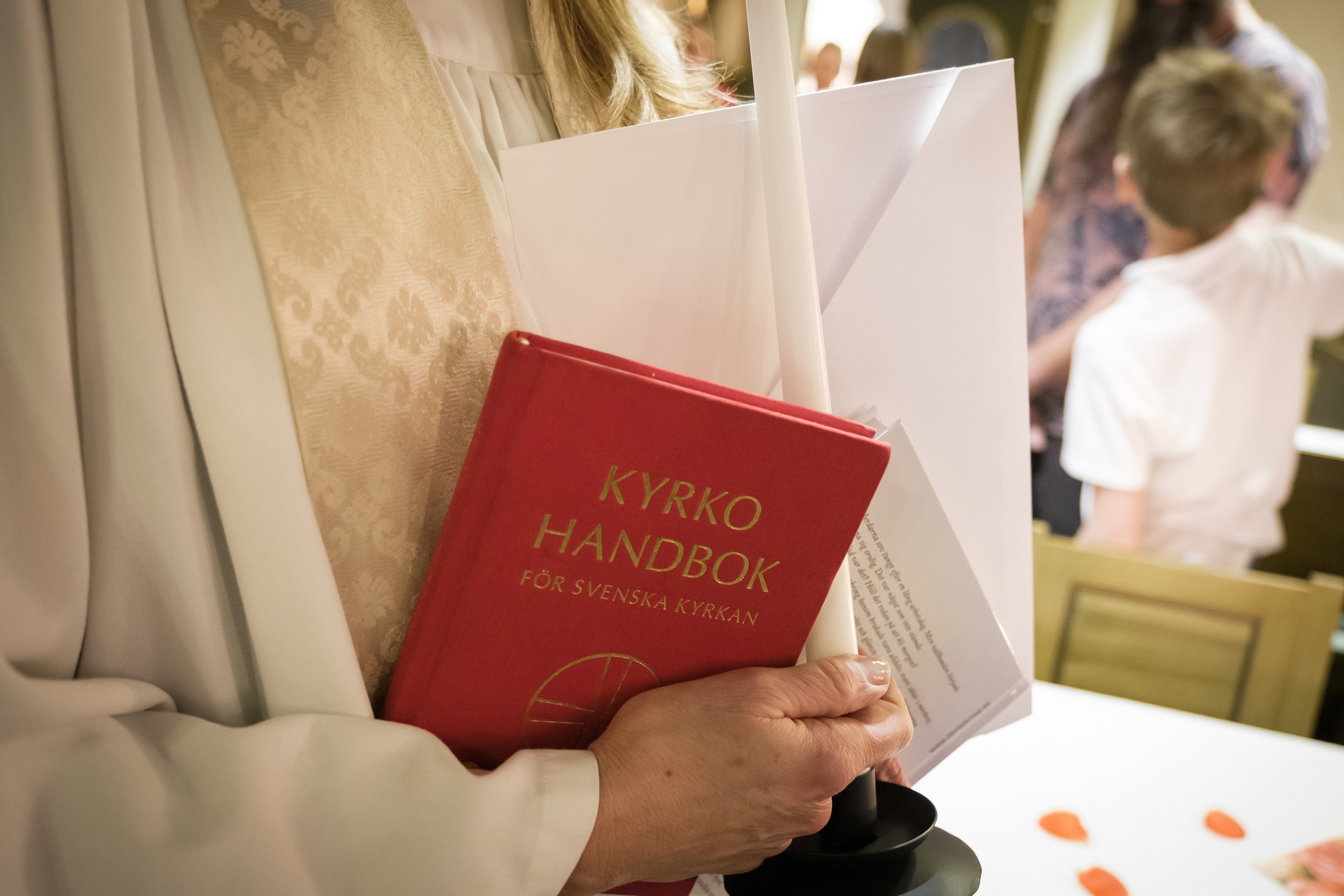 En präst håller ett ljus, ett vitt kuvert och en kyrkohandbok i händerna.
