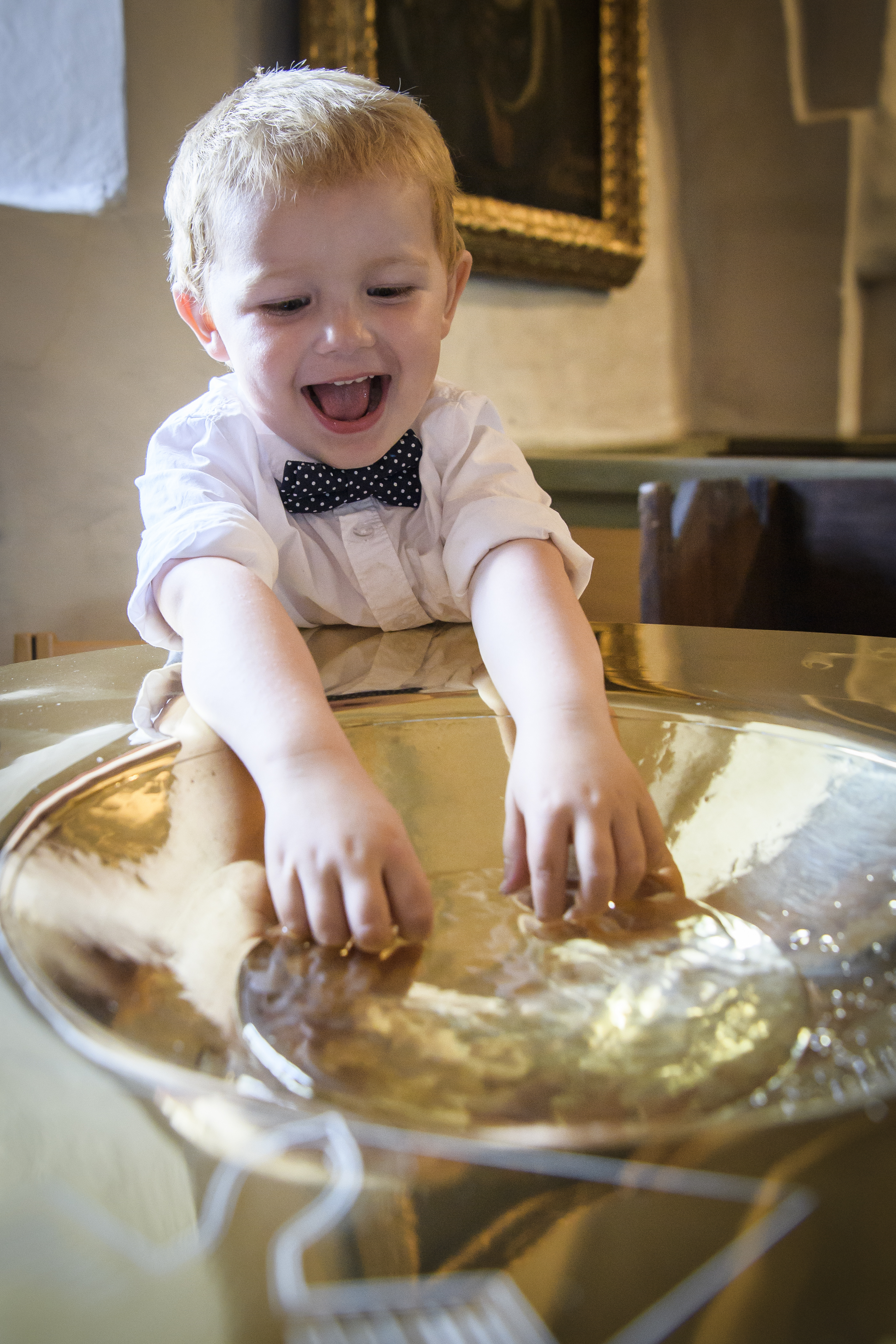 En liten pojke i vit skjorta och prickig fluga doppar sina fingrar i dopfuntens vatten.