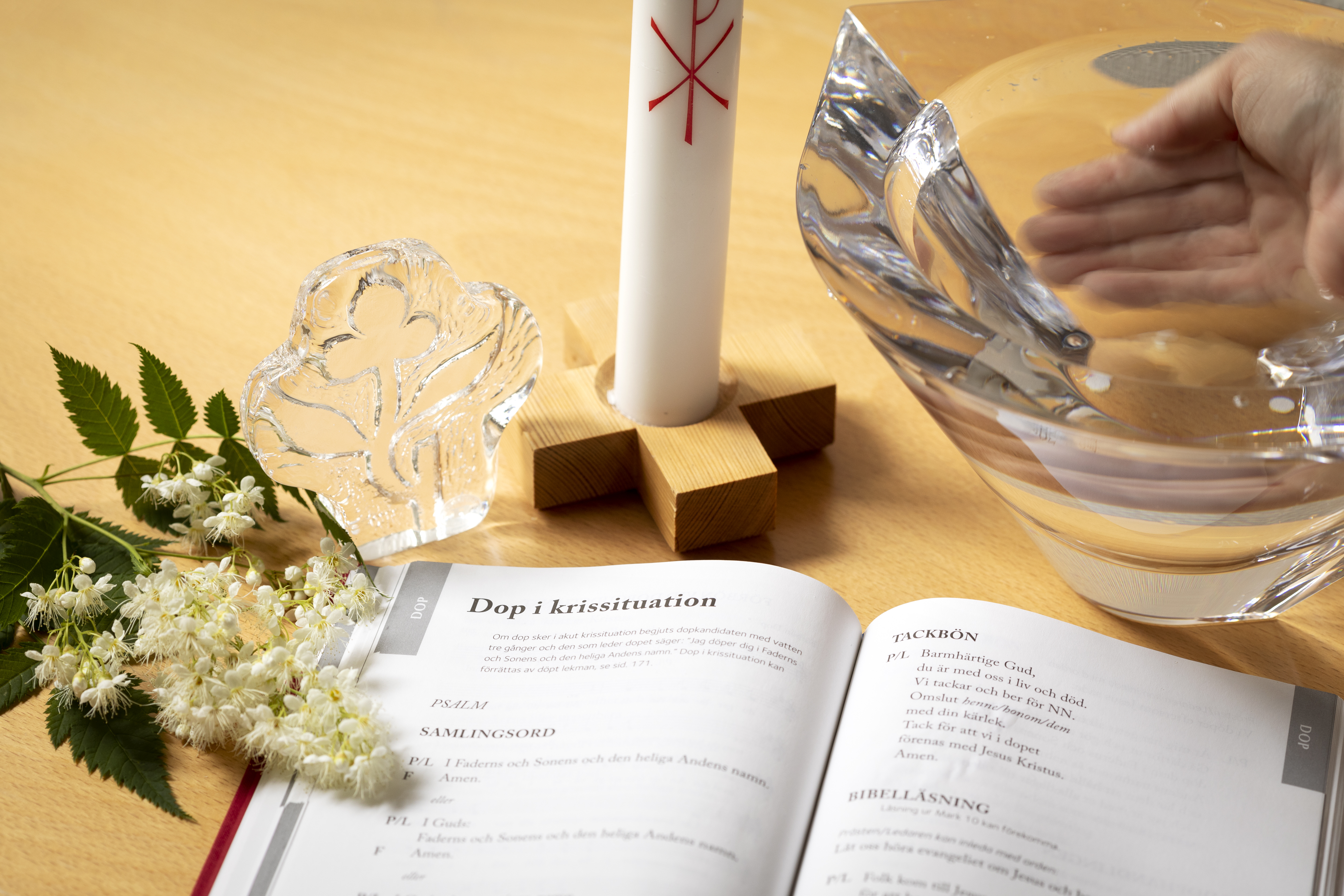 Bredvid en glasskål och ett dopljus ligger en uppslagen bok med rubriken Dop i krissituation.