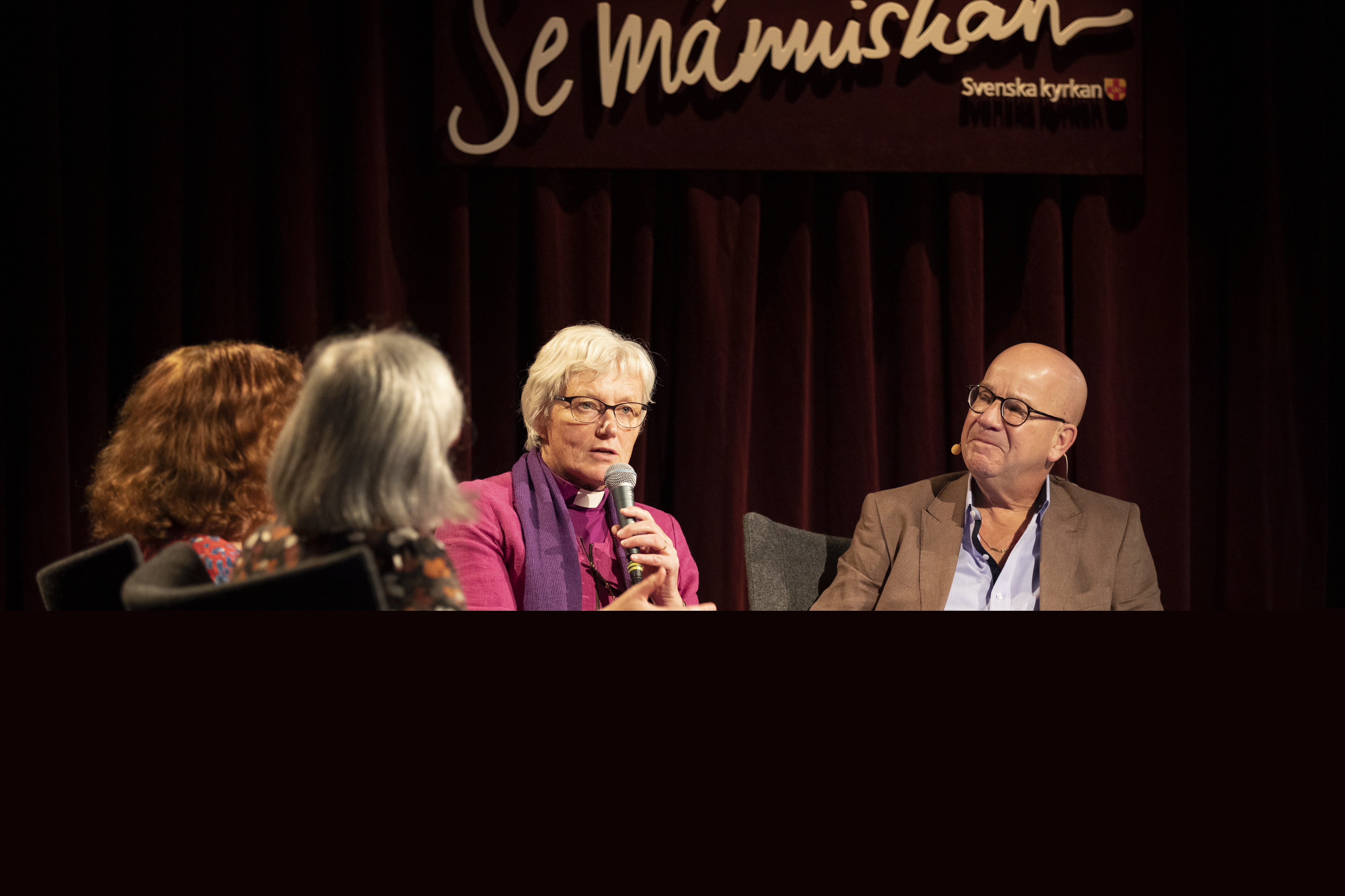 Ärkebiskop Antje Jackelén sitter i en studiomiljö och pratar i en mikrofon. På väggen bakom står det Se människan.