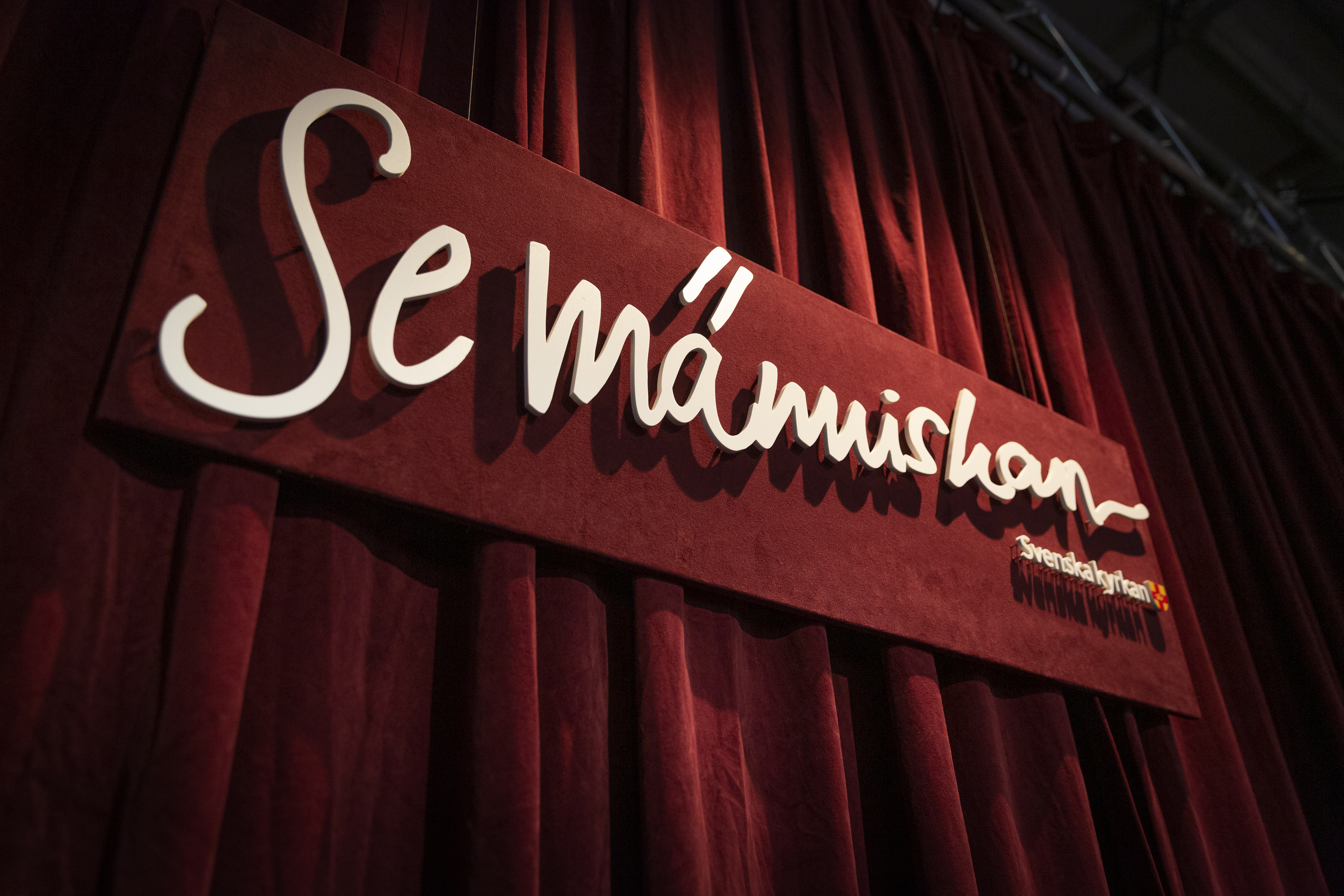 Framför ett rött draperi hänger en skylt med texten Se människan.