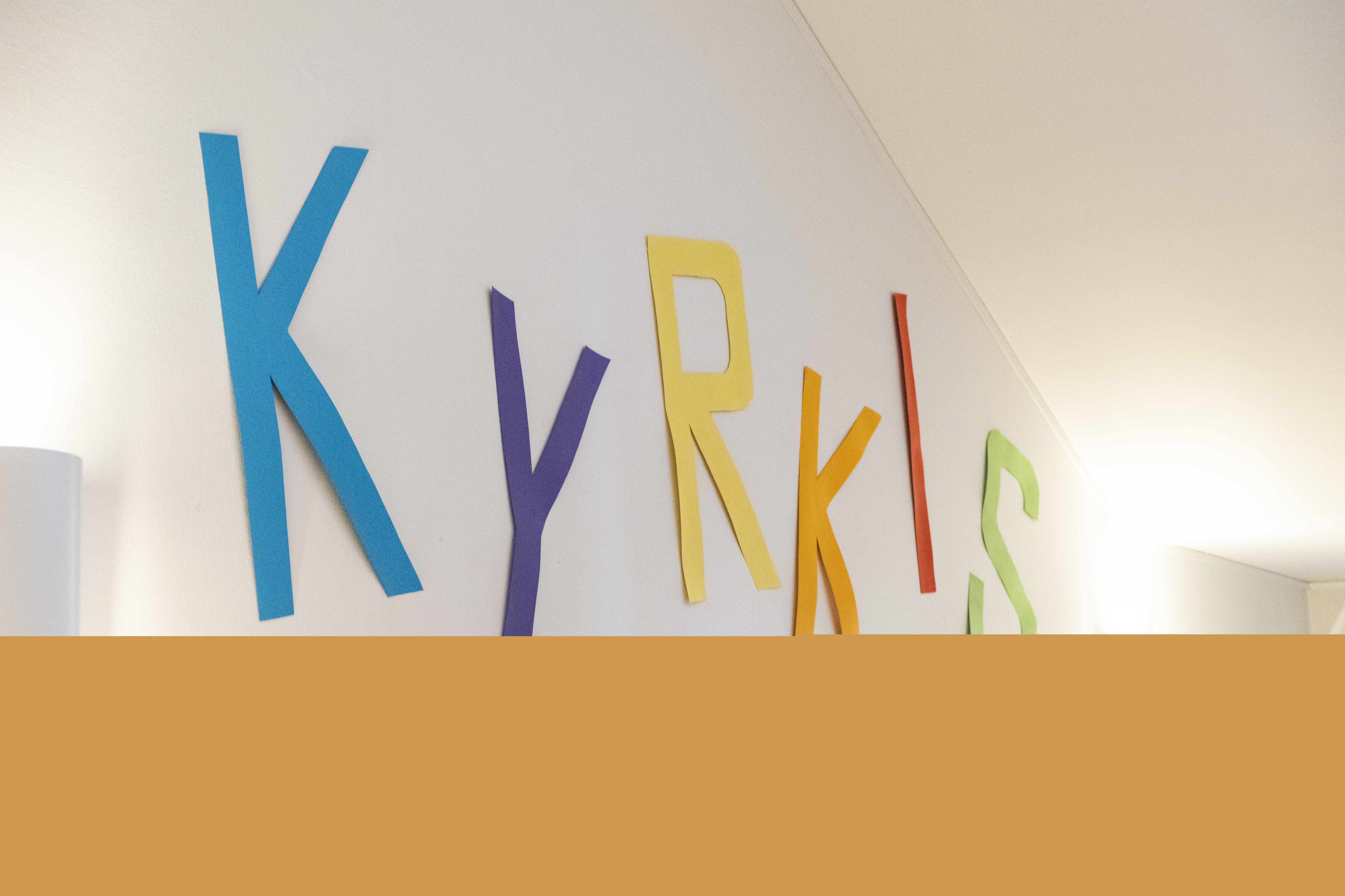 Ordet "KYRKIS" sitter upp på en vit vägg. Bokstäverna är urklippta ur färgat papper i blått, lila, gult, rött och grönt.