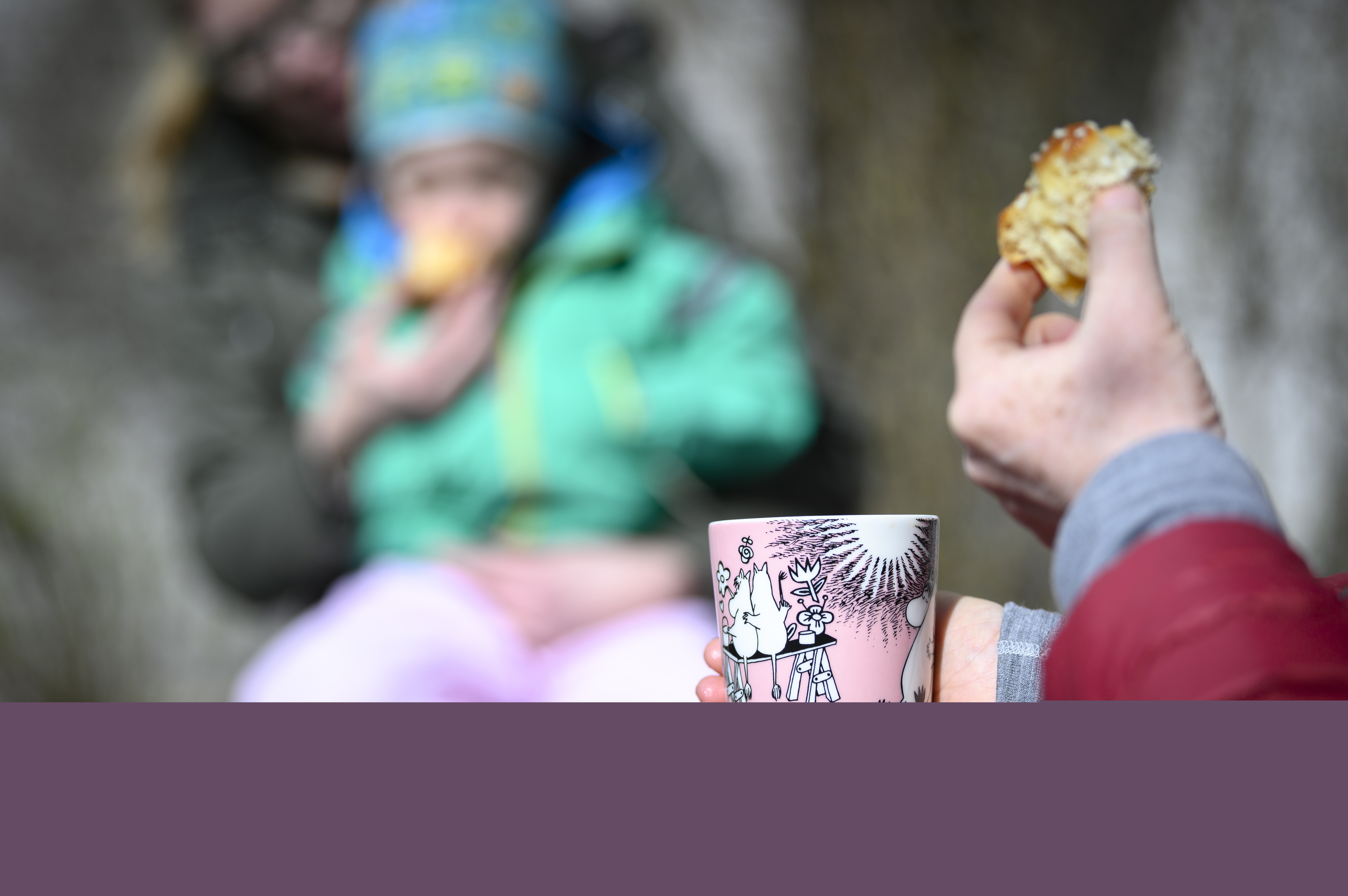 Någon sitter och fikar kaffe och bulle. I bakgrunden sitter ett barn i knät på en vuxen.