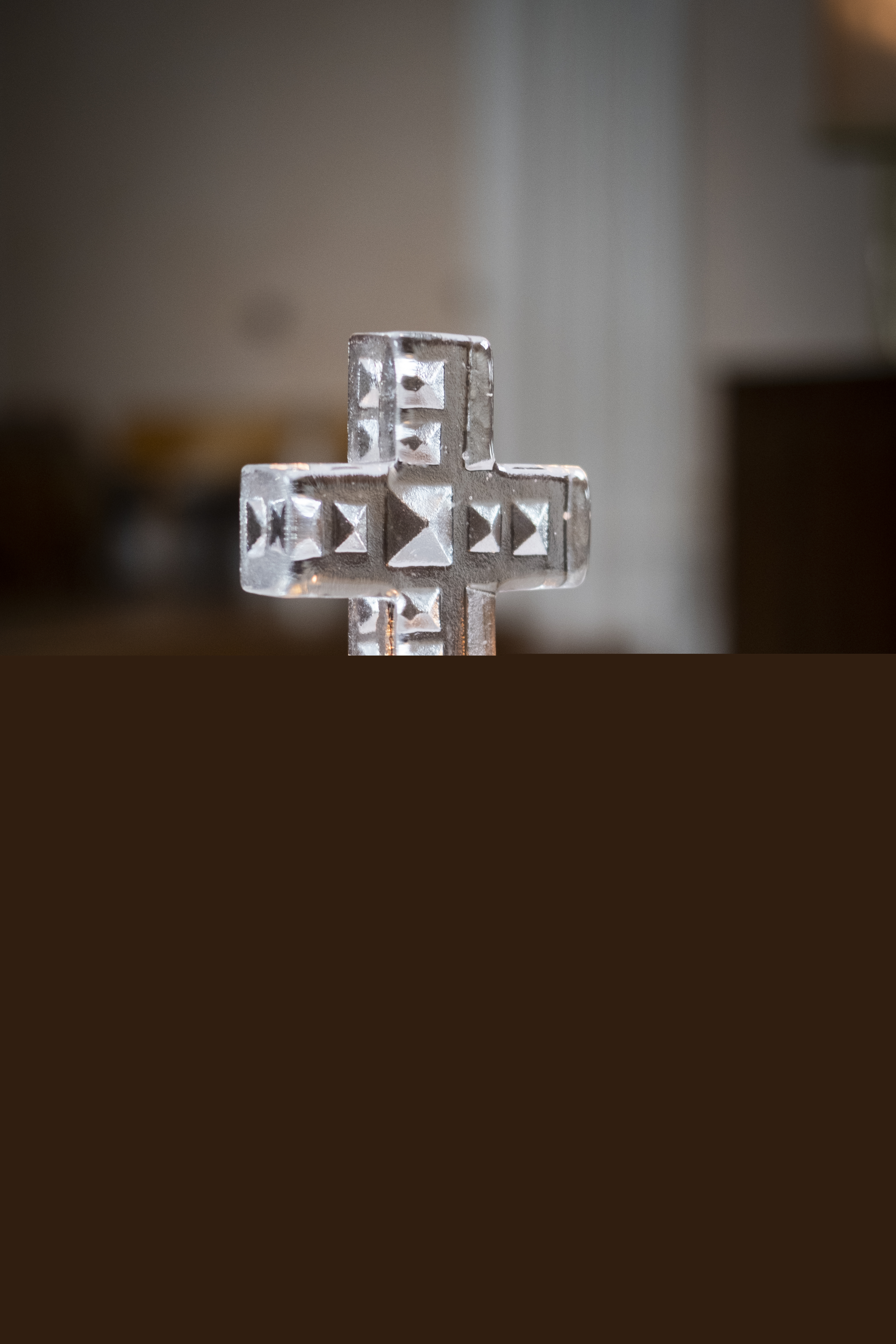 Ett kors av handgjort glas står på ett altare. Framför korset brinner ett ljus på en trefotad ljusstake.