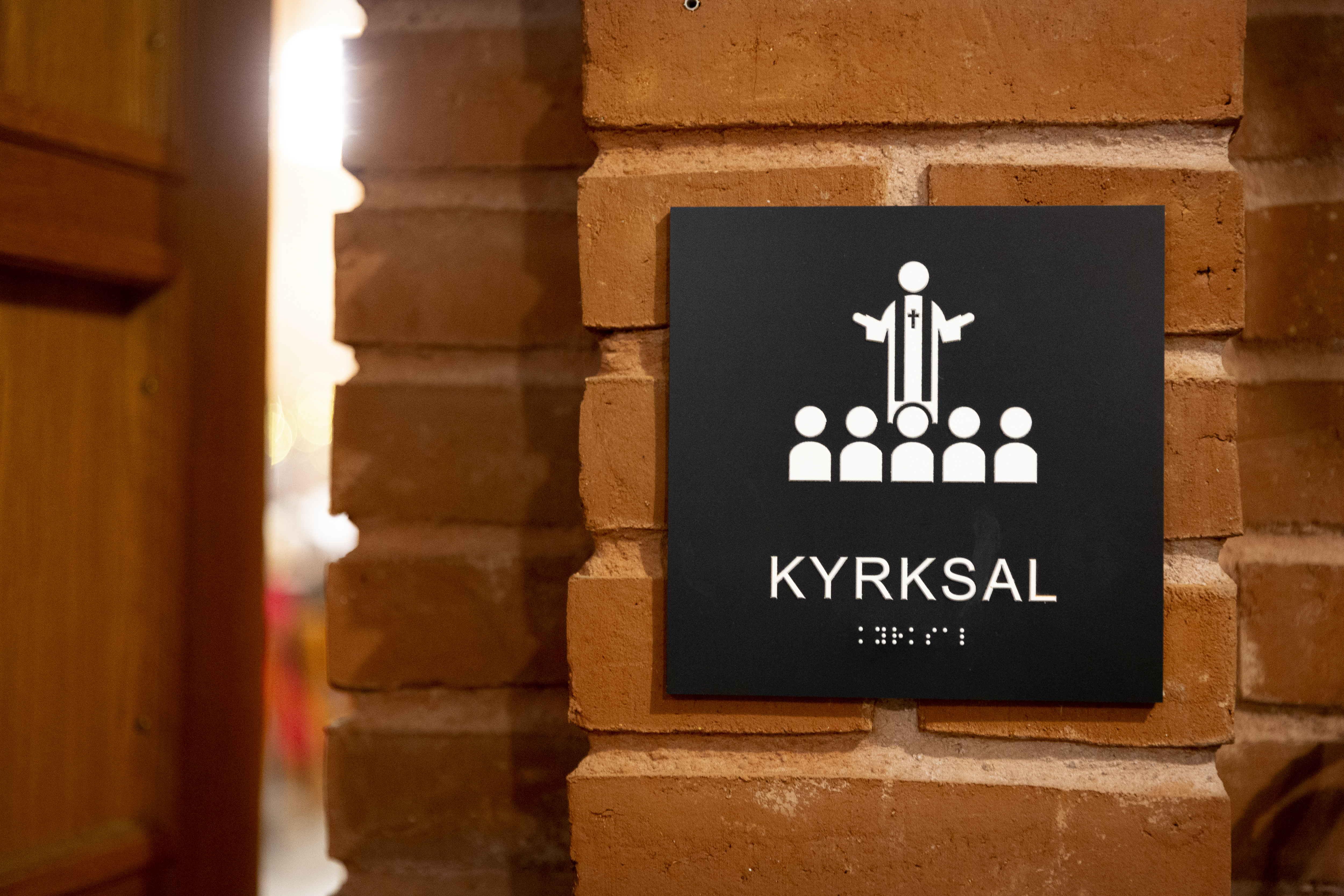 En skylt med texten och blindskriften Kyrksal sitter på en tegelvägg bredvid dörren.