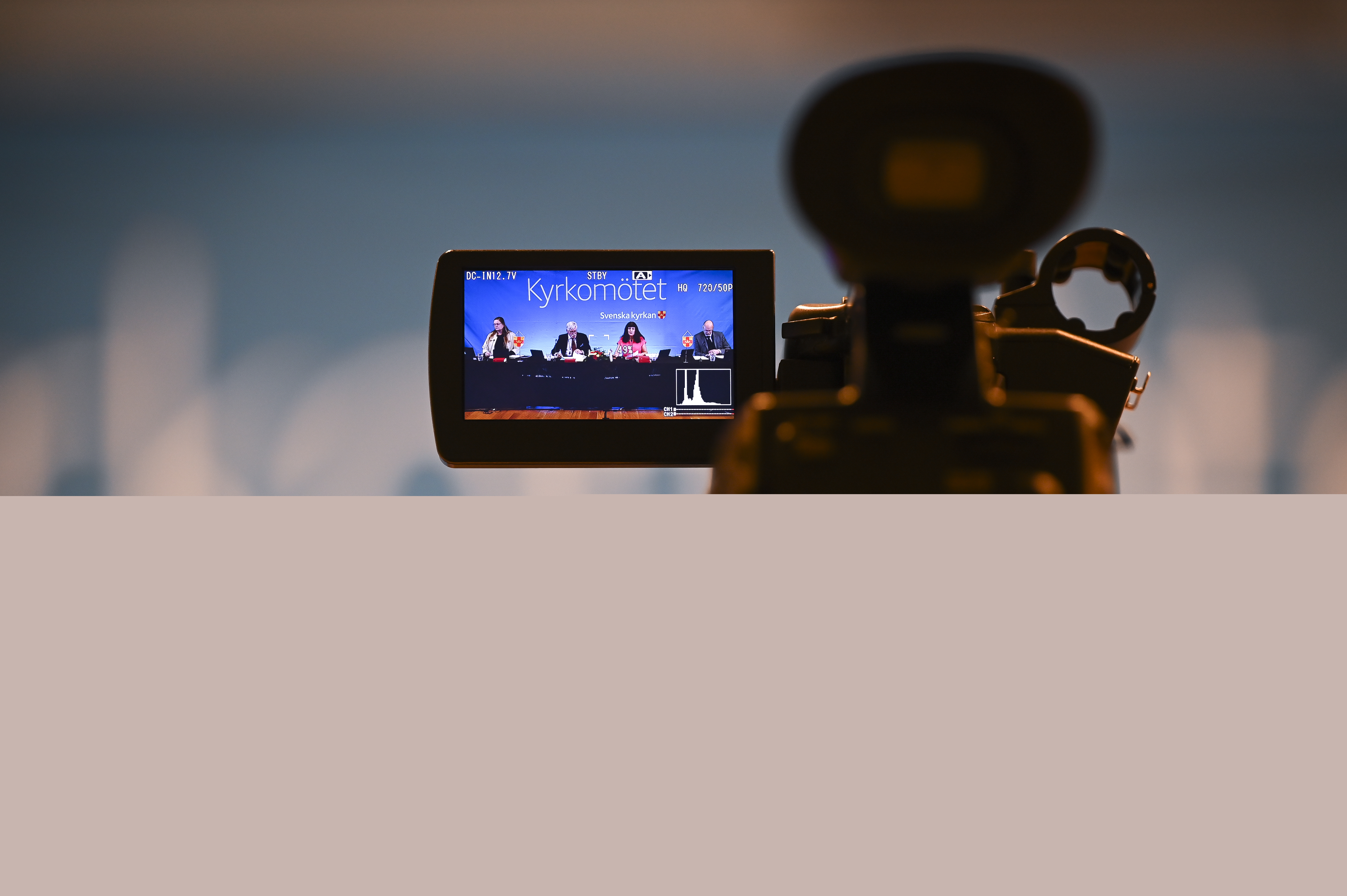 På skärmen till en videokamera syns en panel med fyra personer framför en fond med texten Kyrkomötet.
