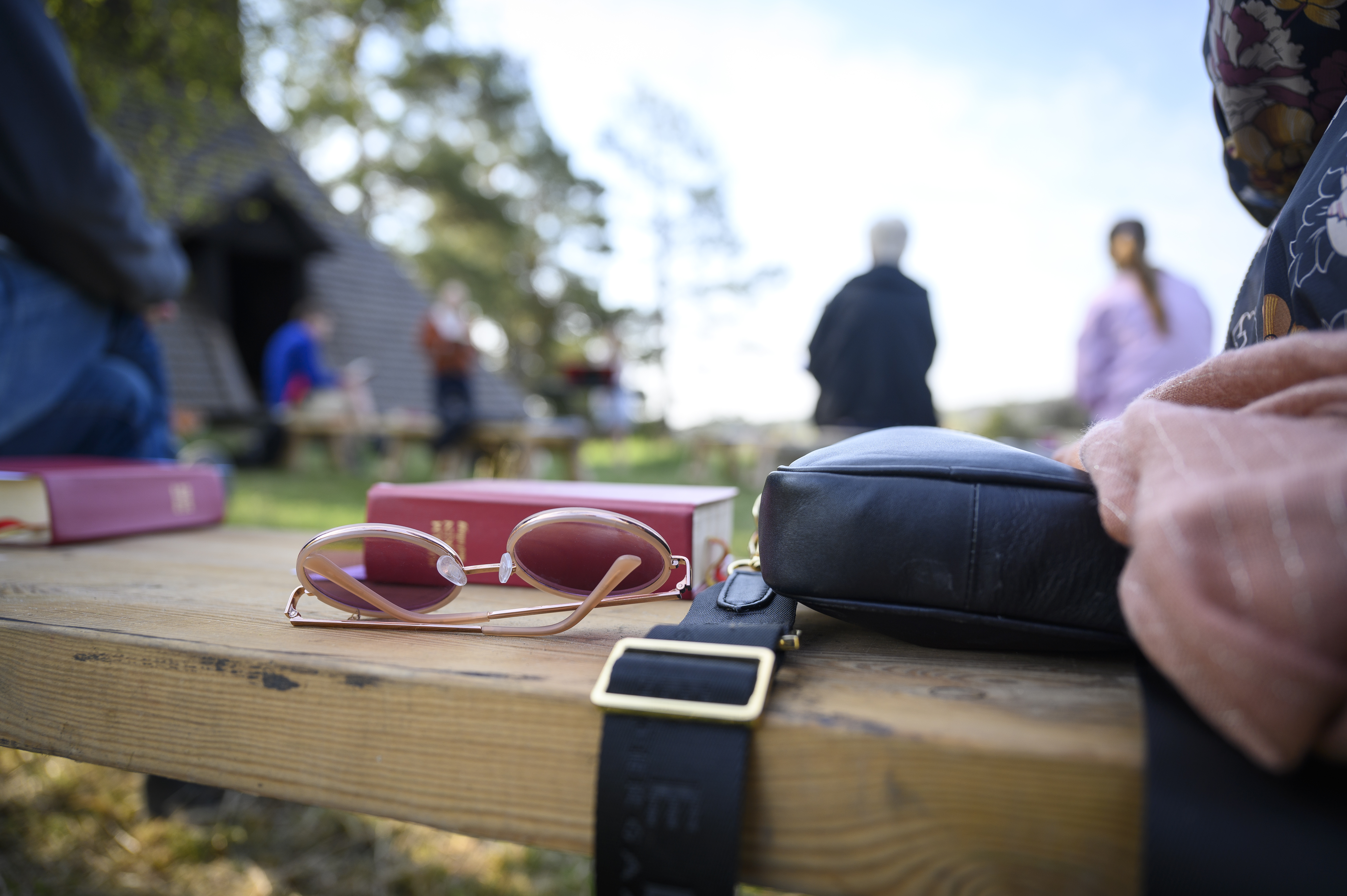 Ett par solglasögon och några psalmböcker ligger på en bänk utomhus. Människor syns i bakgrunden.