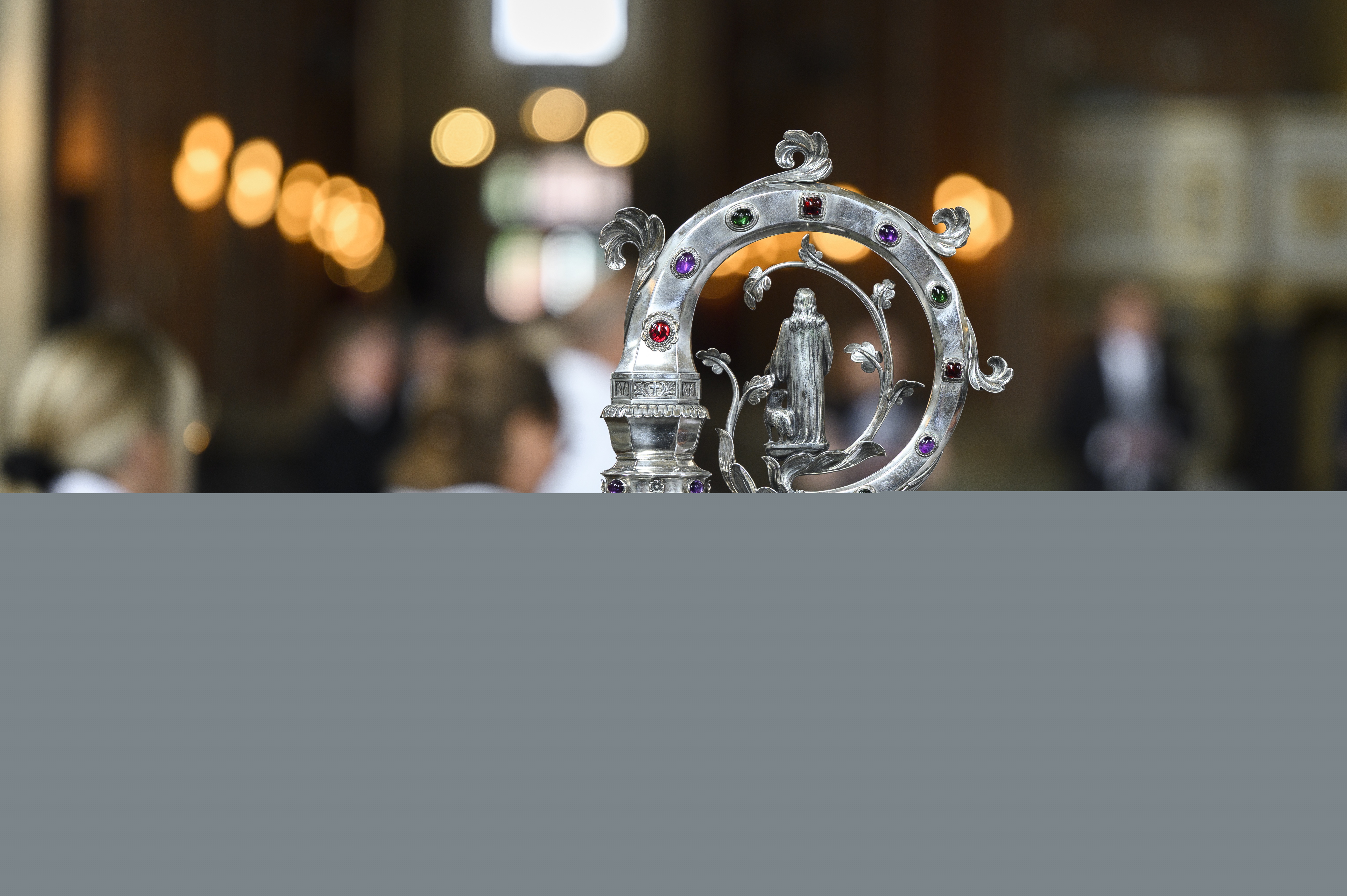 En silvrig biskopskräkla med ädelstenar. I bakgrunden några kvinnor i vita kåpor.