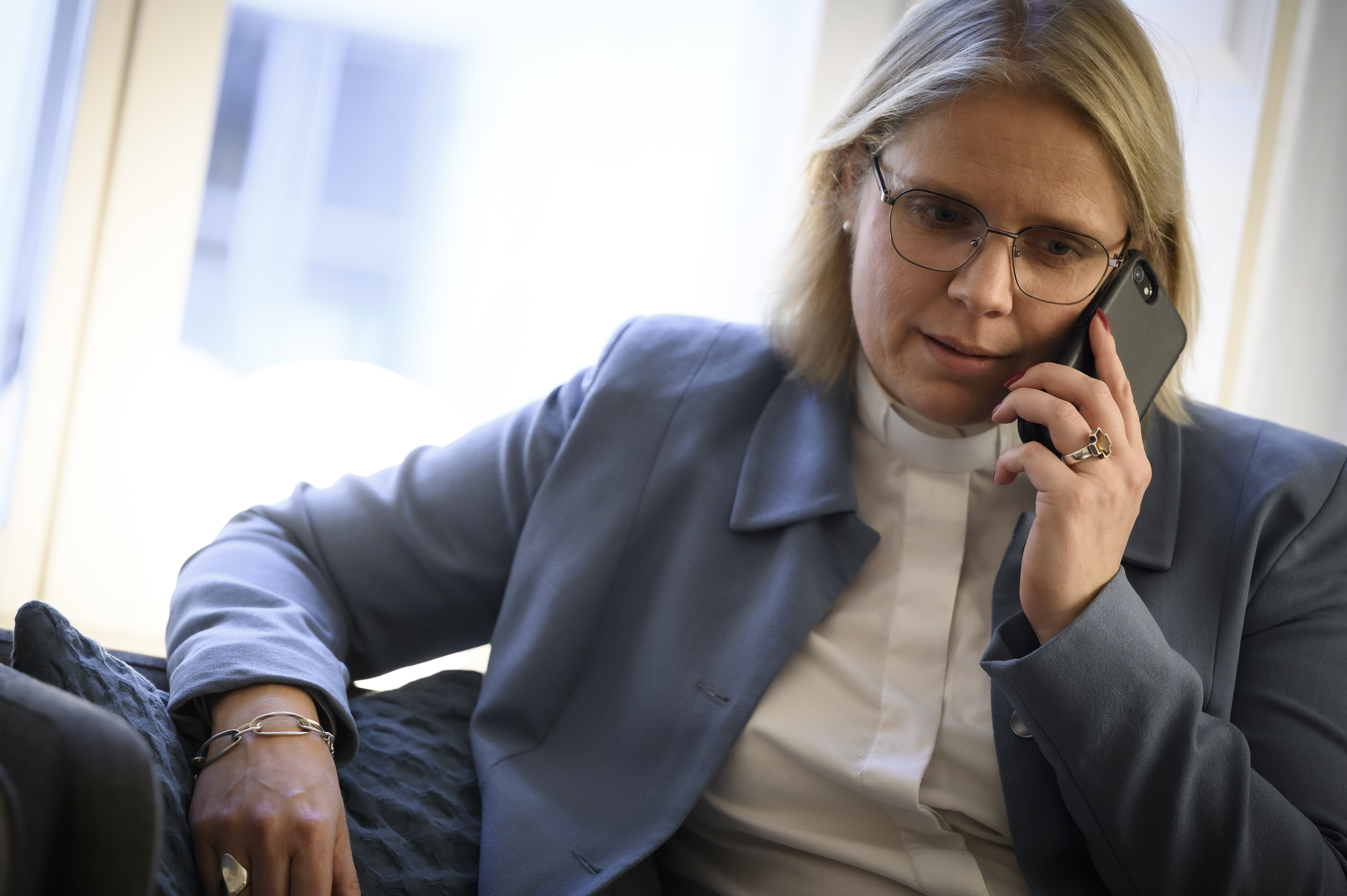 En kvinnlig präst i ljusblå kavaj sitter i en soffa och pratar i telefon.