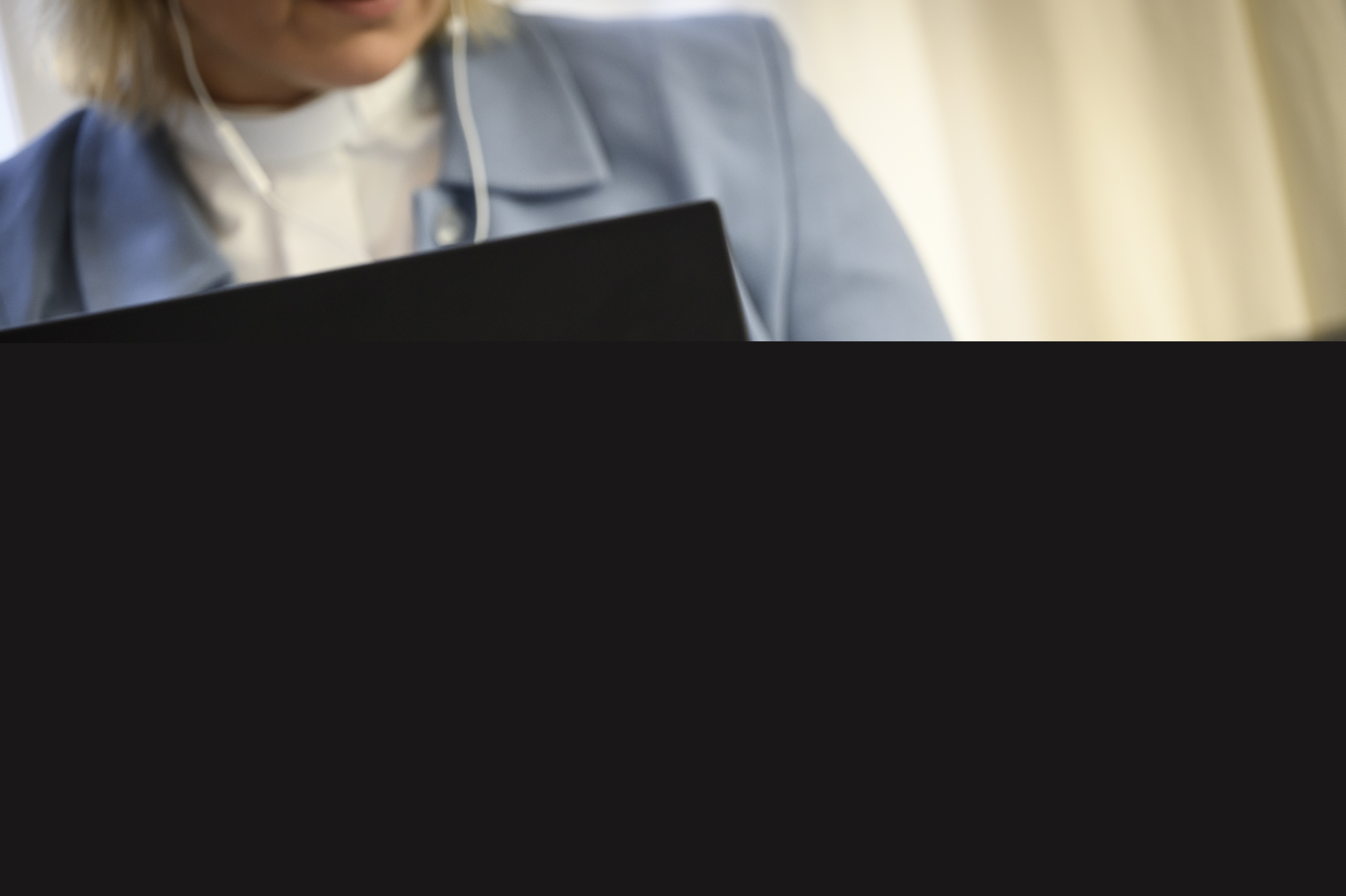 En kvinnlig präst i ljusblå kavaj sitter i en soffa med en laptop i knät och en mobiltelefon i handen.