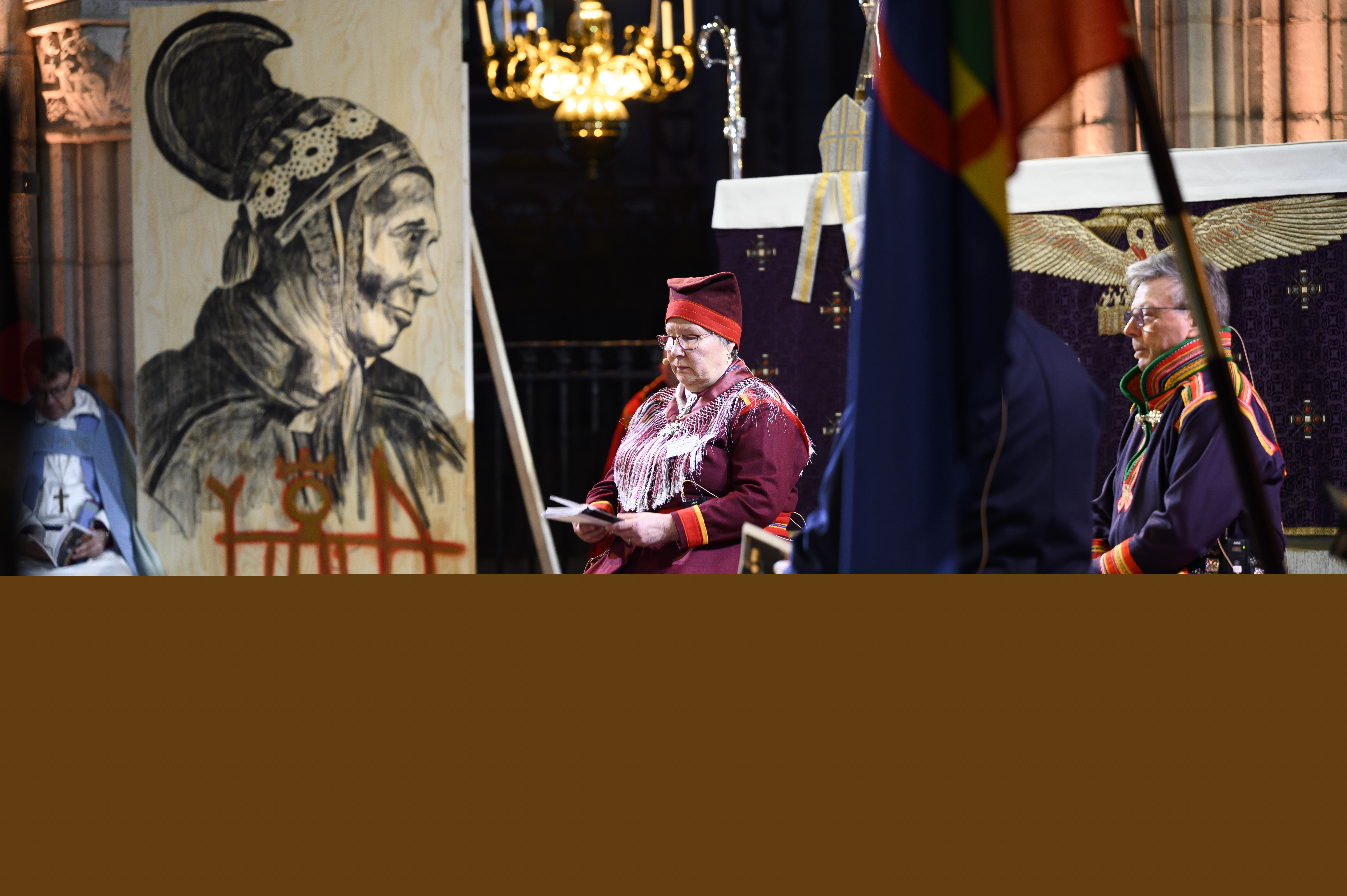 Tre personer i samedräkt sitter på grovhuggna bänkar runt en lägereld framför Uppsala domkyrkas altare.