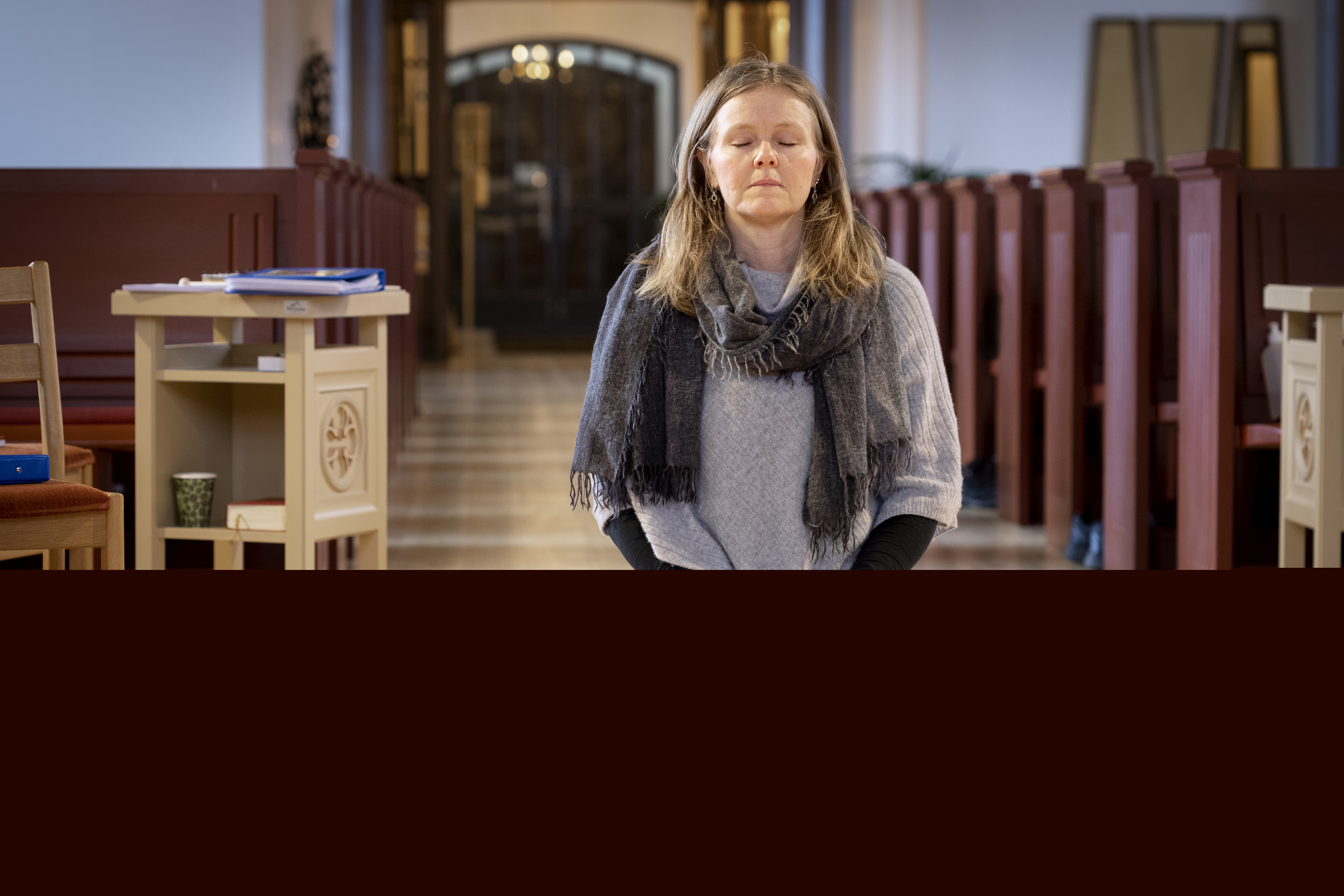 En kvinna sitter på en bönepall på en kyrkas golv, med knäppta händer och slutna ögon.