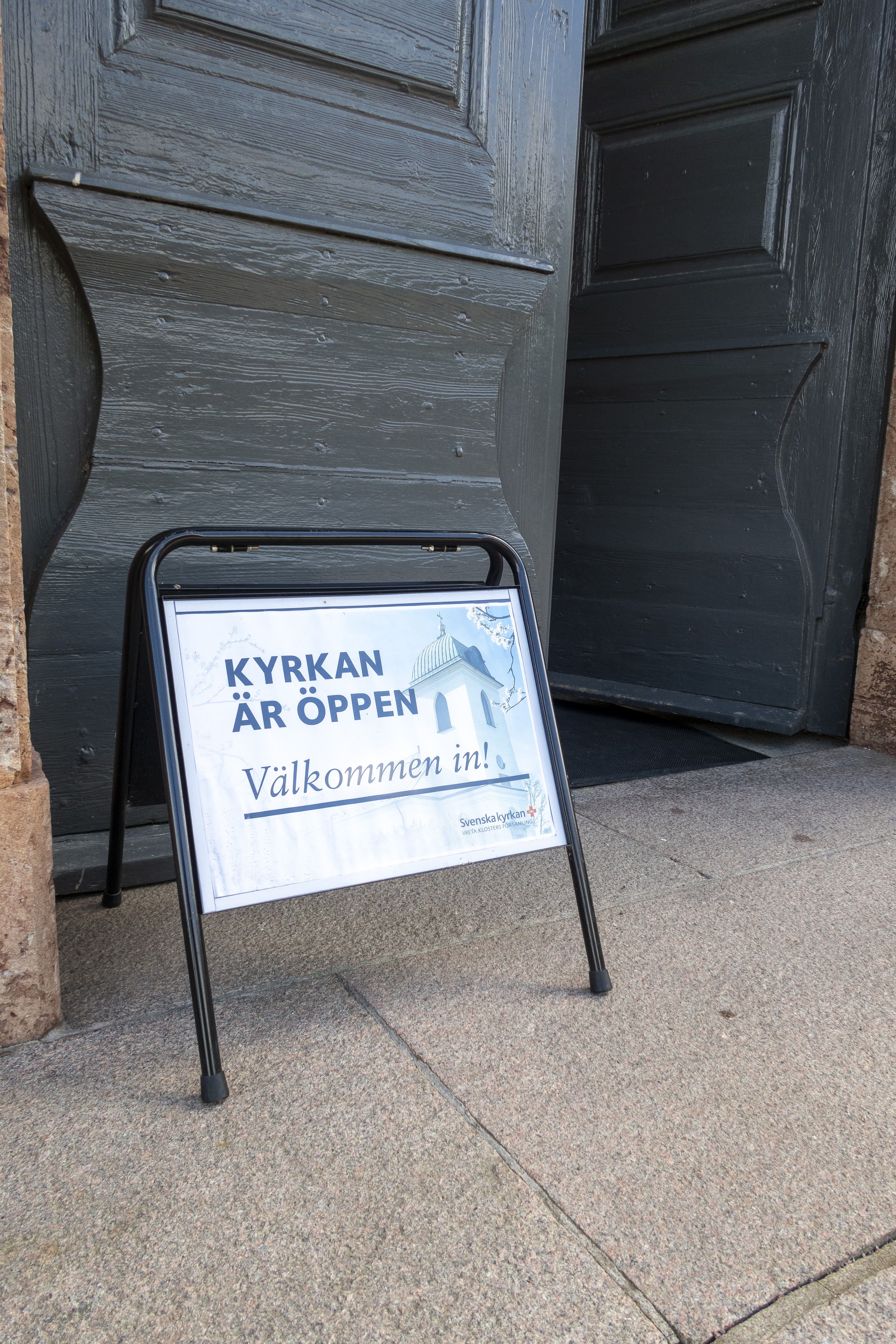 En skylt med texten Kyrkan är öppen välkommen in, står utanför en port.