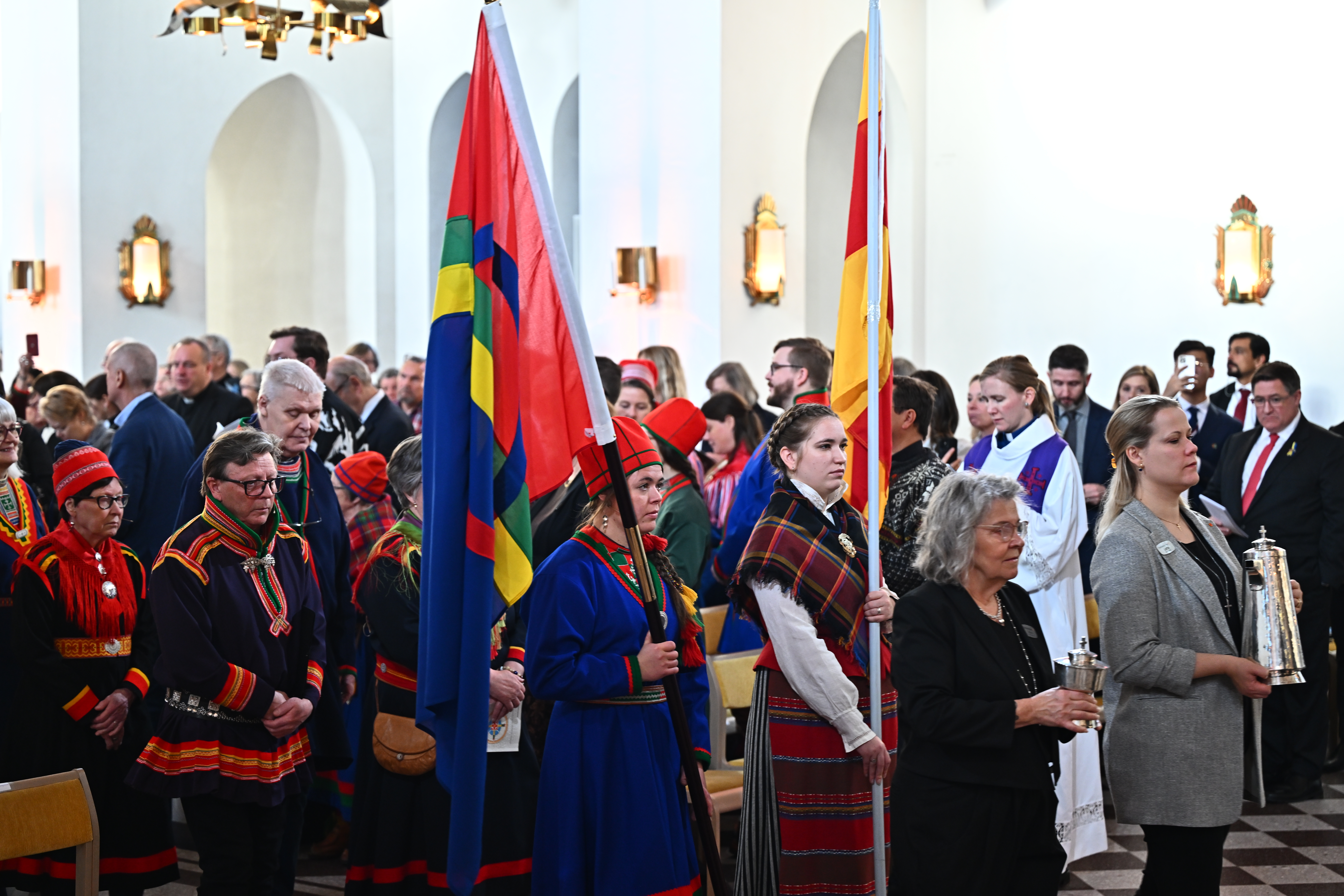Två kvinnor i samekläder står i en kyrka med varsin flagga, den samiska och Svenska kyrkans flagga. Många personer i bakgrunden.