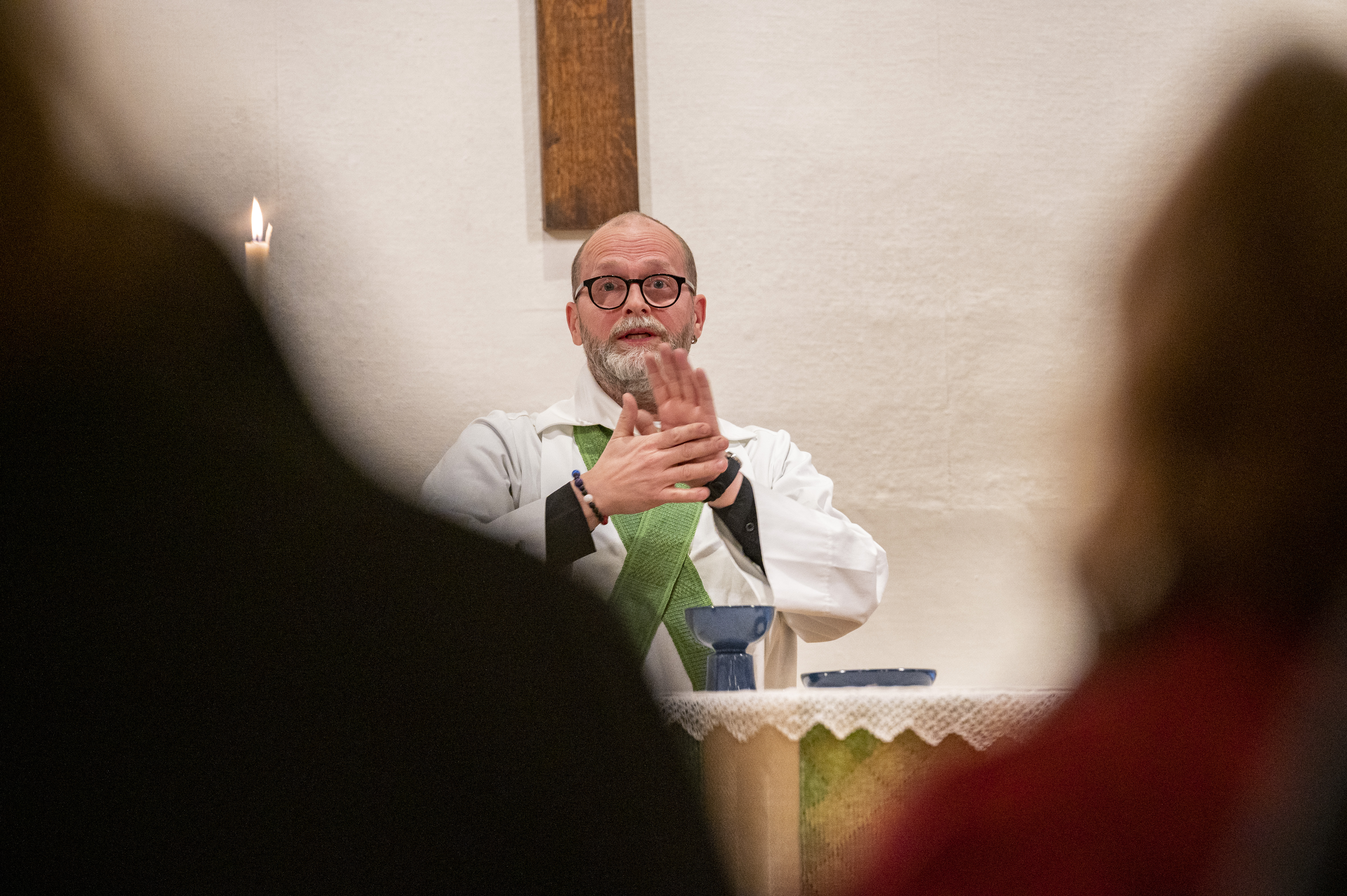 En manlig präst står vid altaret och gestikulerar med händerna. Suddiga personer i förgrunden.