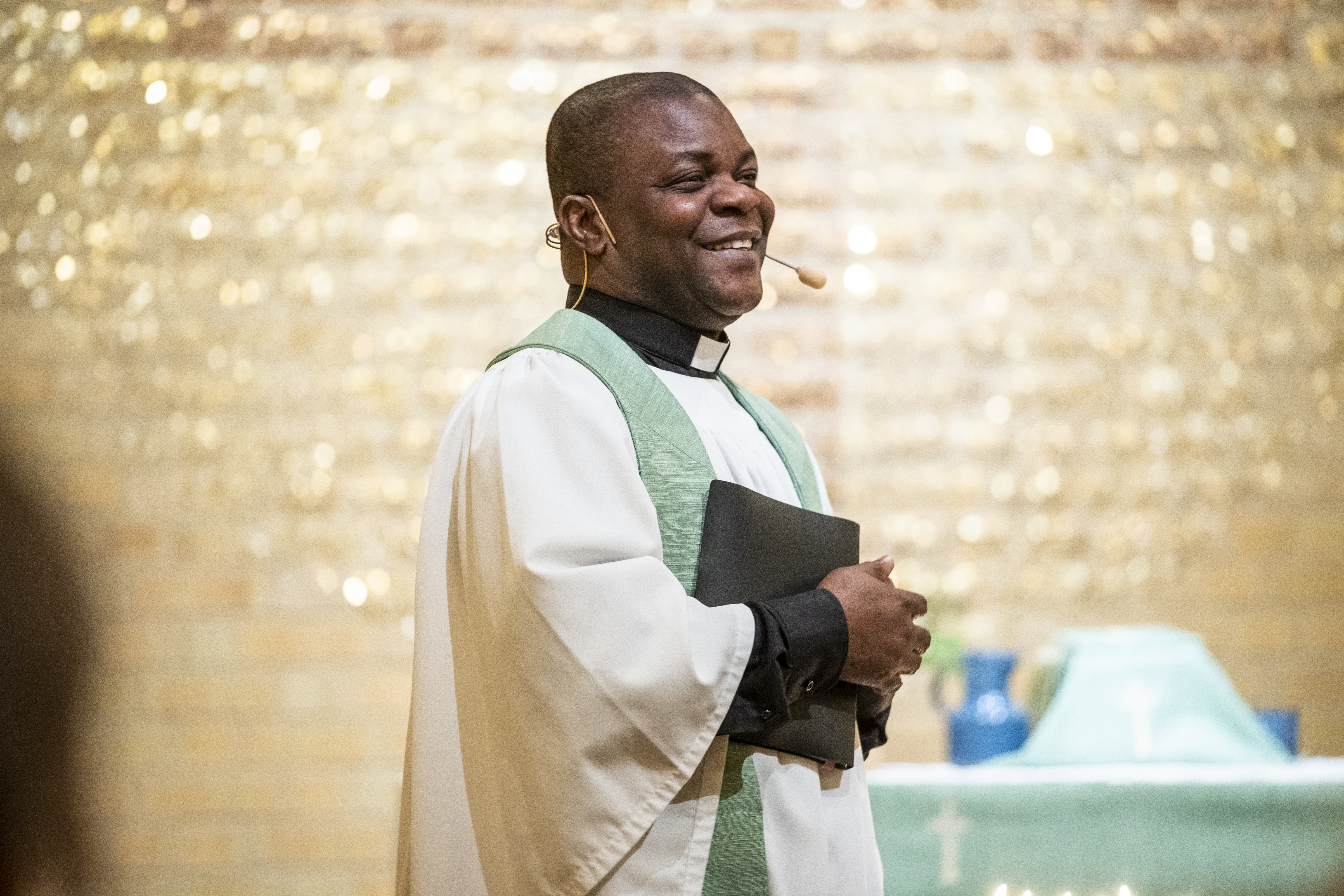 En manlig präst håller i en gudstjänst. Han har en svart bok i famnen och ler stort.