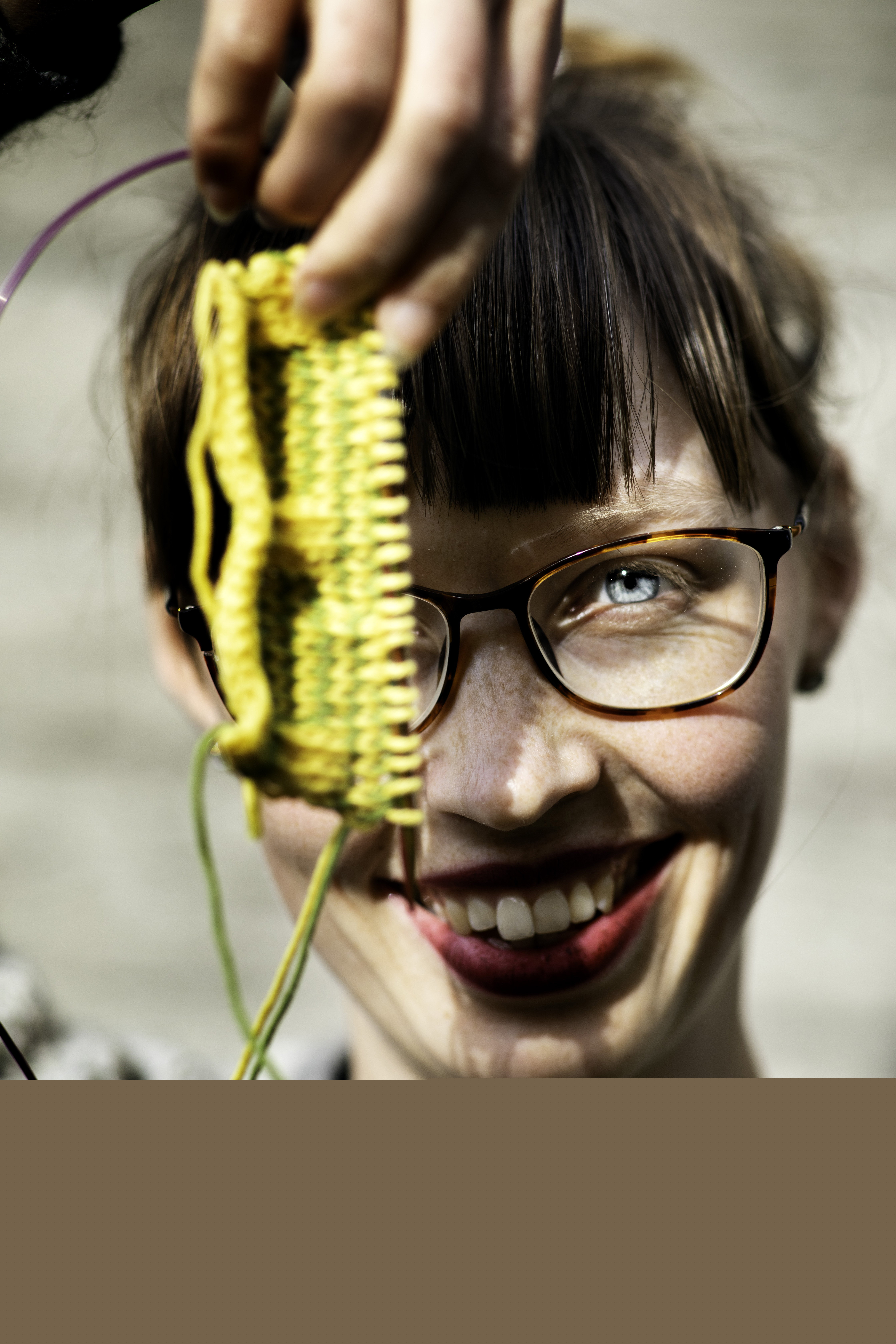 En kvinna med lugg och rött läppstift håller upp en gul stickning mot kameran och ler stort.