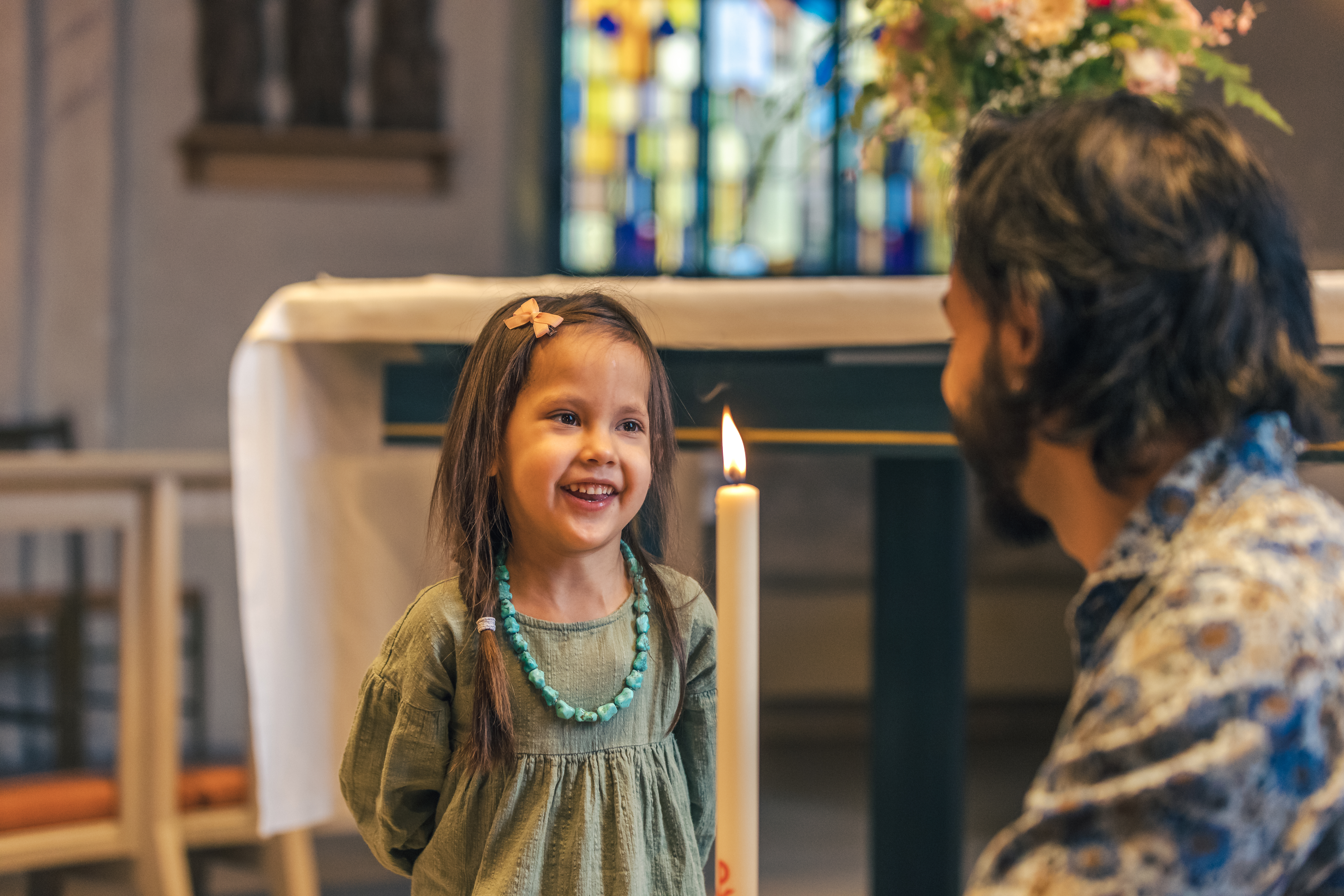 En man sitter på huk i en kyrka och håller i ett dopljus. En liten flicka står framför och ler stort.