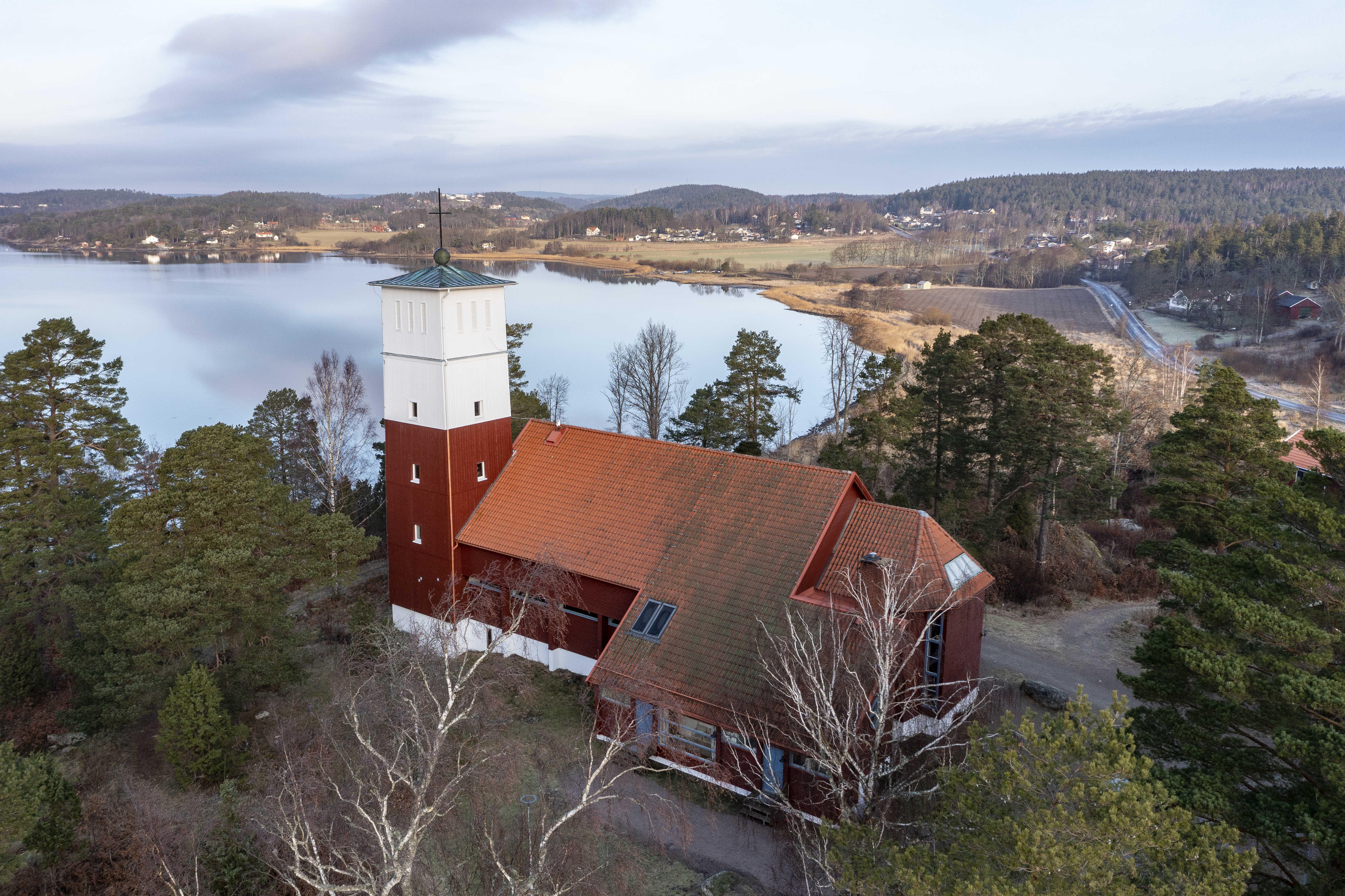 Drönarbild på en faluröd kyrka med vitt torn tegeltak, vackert belägen vid en sjö.