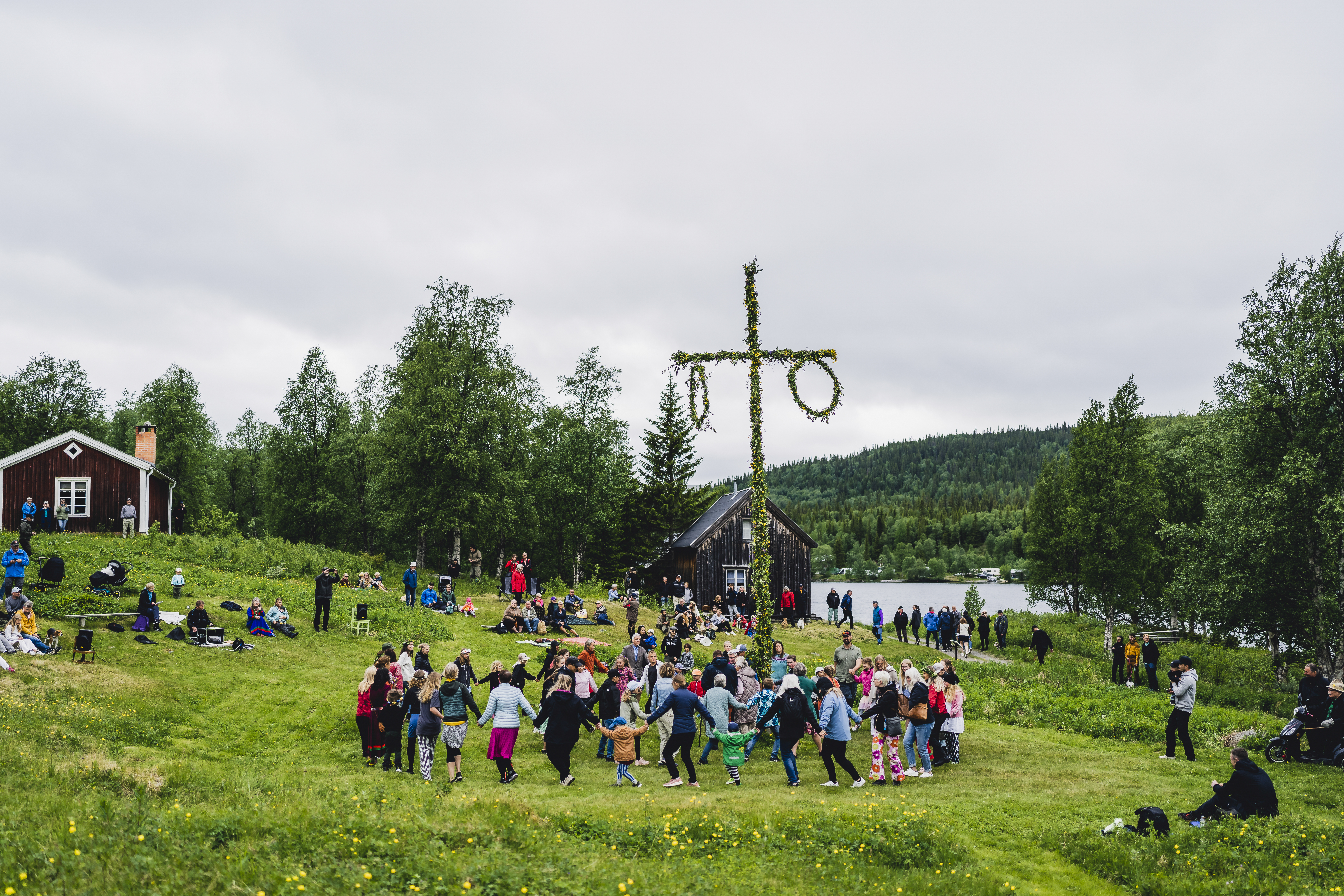 Ett midsommarfirande vid en sjö. Vuxna och barn dansar runt stången.