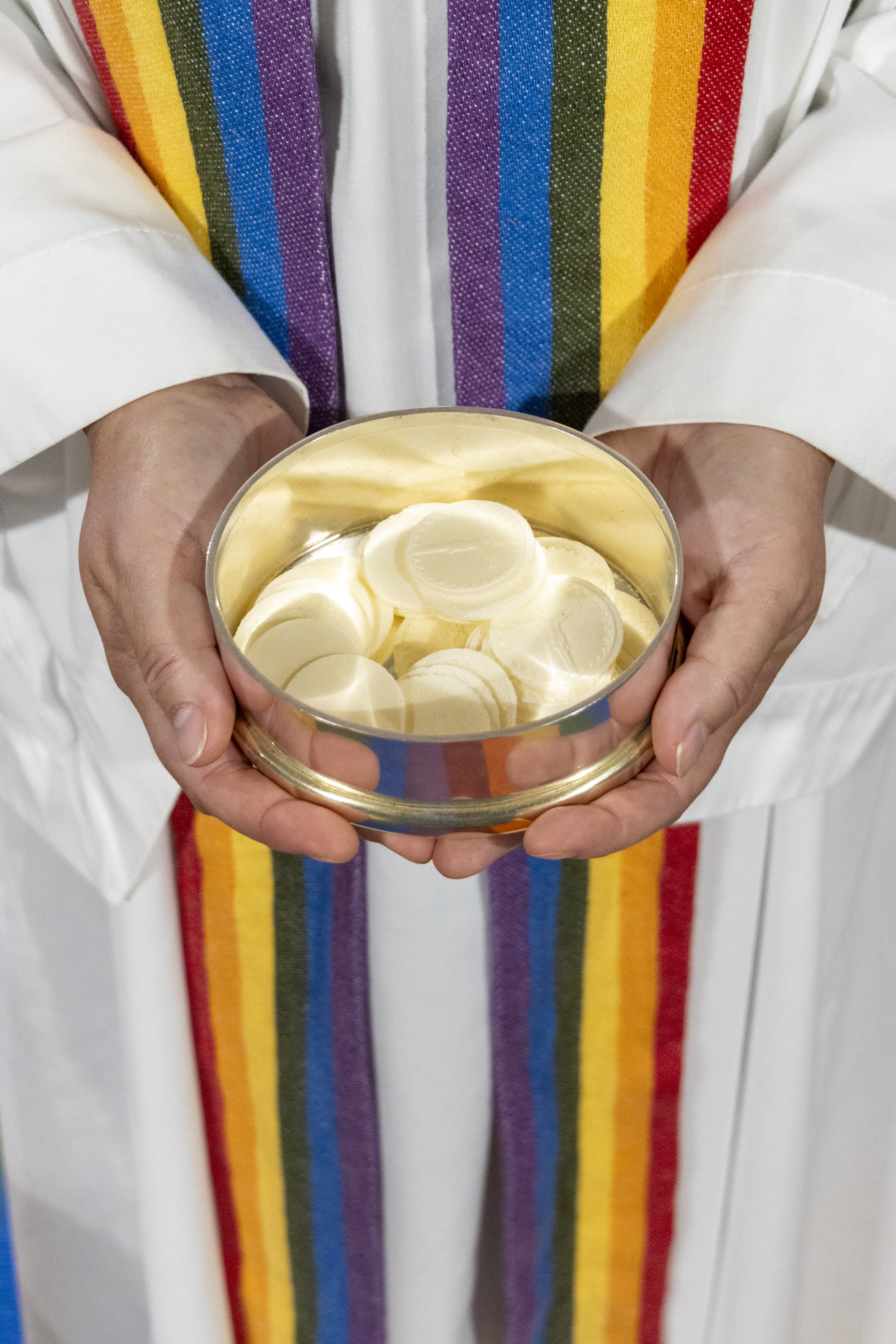 En präst med en regnbågsfärgad stola håller i en skål oblat.