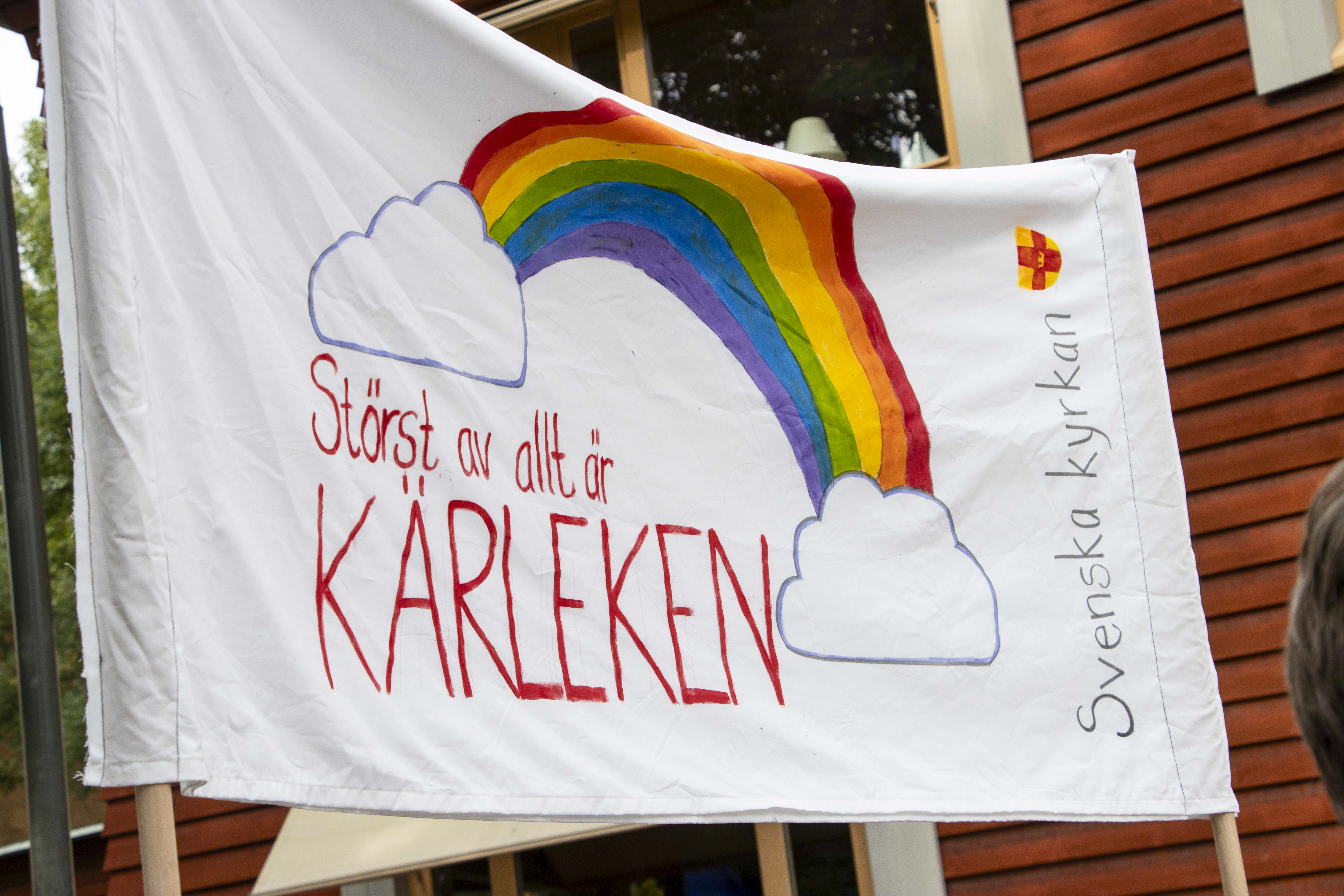En tygskylt med regnbågsflaggan, Svenska kyrkans logotyp och texten ”störst av allt är kärleken”.